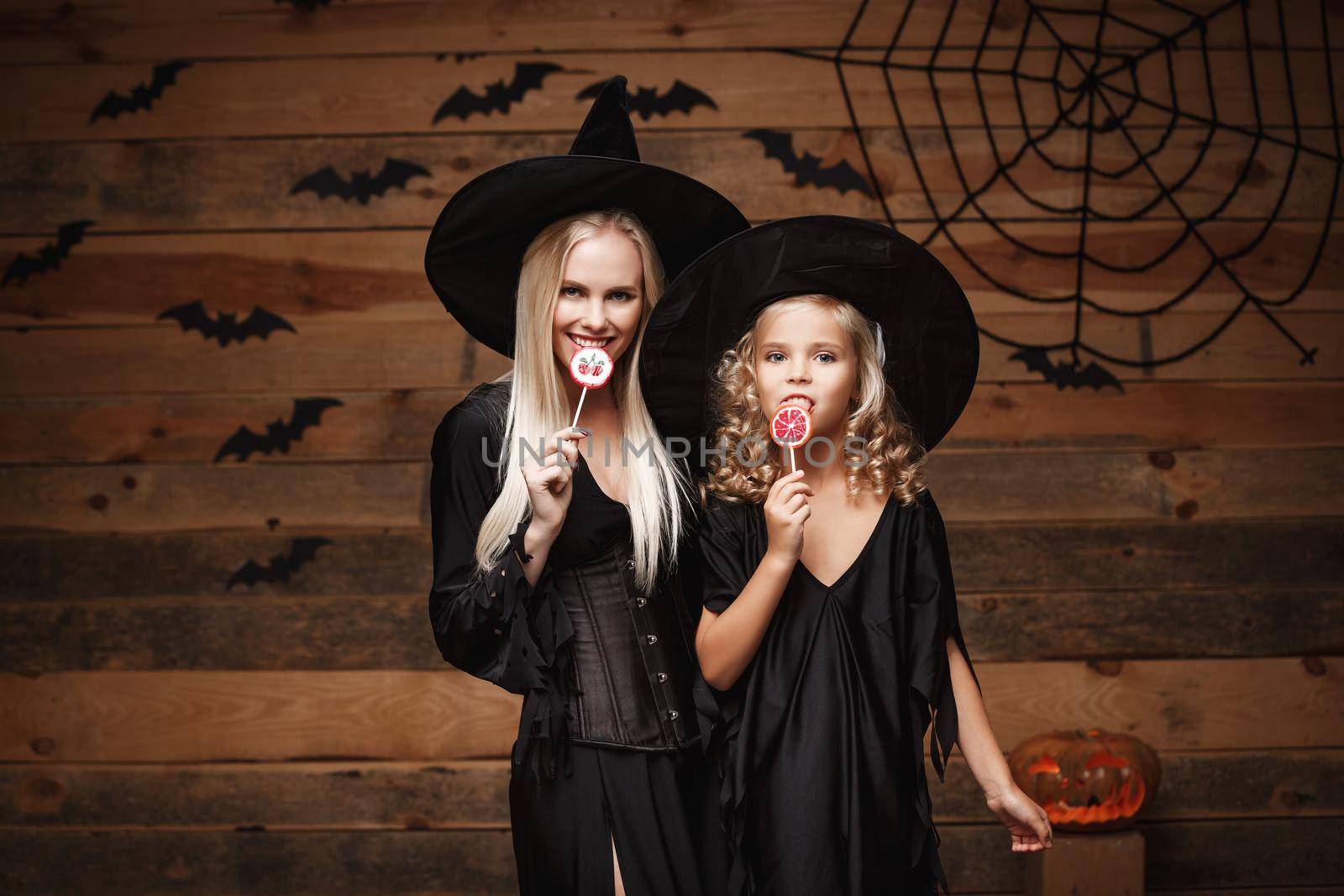 Halloween Concept: beautiful caucasian mother and her daughter in witch costumes celebrating Halloween with Halloween candy and sweet over bats and spider web on Wooden studio background