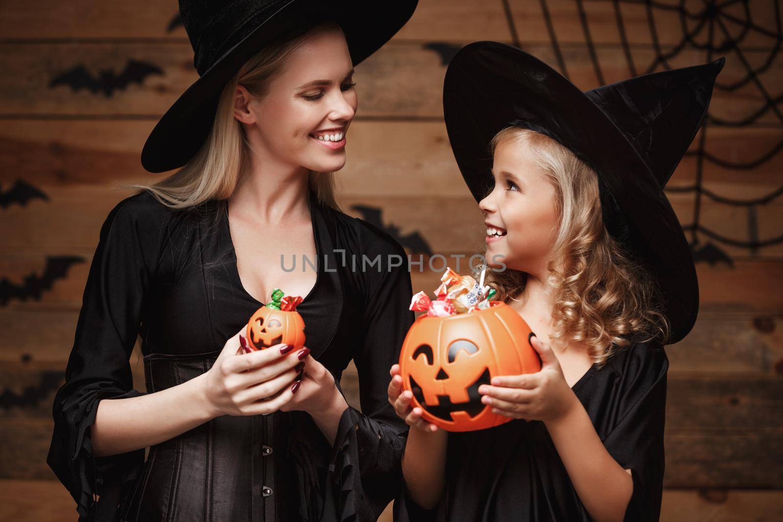 Halloween Concept: beautiful caucasian mother and her daughter in witch costumes celebrating Halloween with sharing Halloween candy and sweet over bats and spider web on Wooden studio background