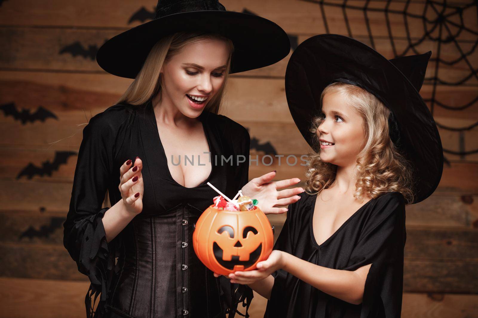 Halloween Concept - beautiful caucasian mother and her daughter in witch costumes celebrating Halloween with sharing Halloween candy and sweet over bats and spider web on Wooden studio background. by Benzoix