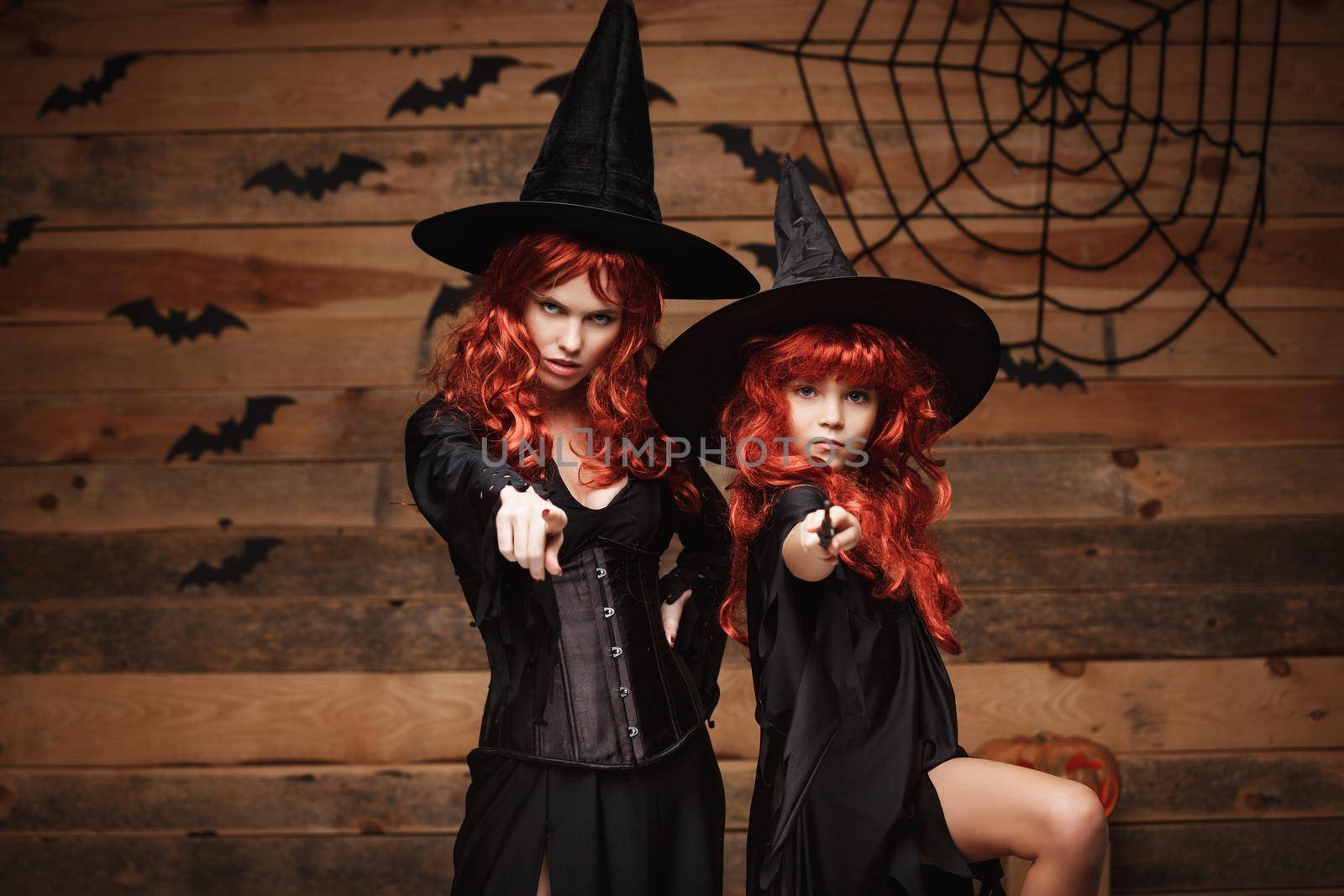 Halloween Concept - Beautiful caucasian mother and her daughter with long red hair in witch costumes and magic wand celebrating Halloween posing with over bats and spider web on Wooden studio background.