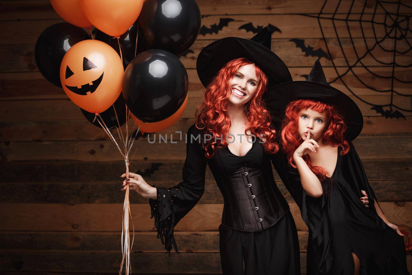 cheerful mother and her daughter in witch costumes celebrating Halloween posing with orange and black balloon over bats and spider web on Wooden studio background