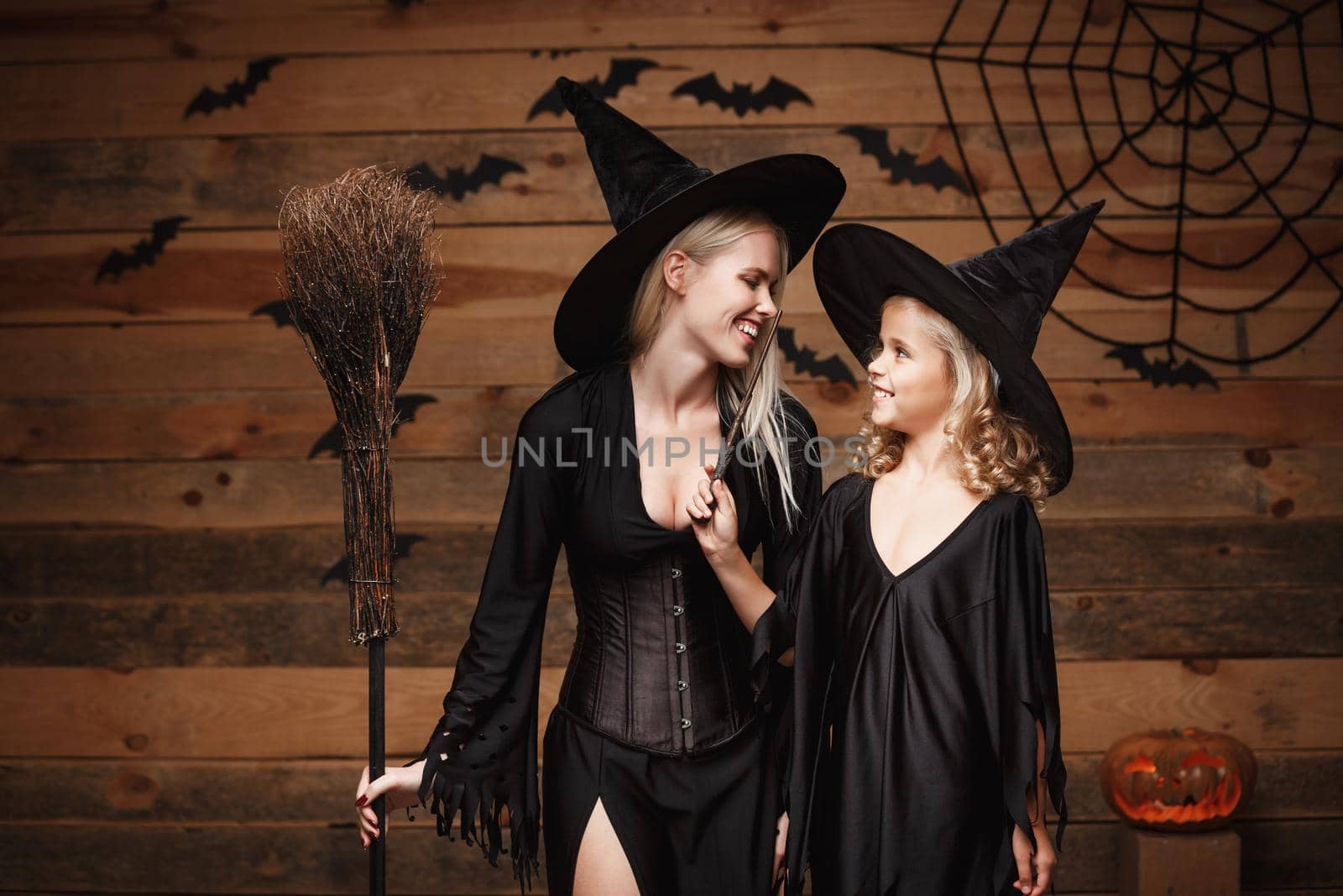 Halloween Concept - cheerful mother and her daughter in witch costumes celebrating Halloween posing with curved pumpkins over bats and spider web on Wooden studio background