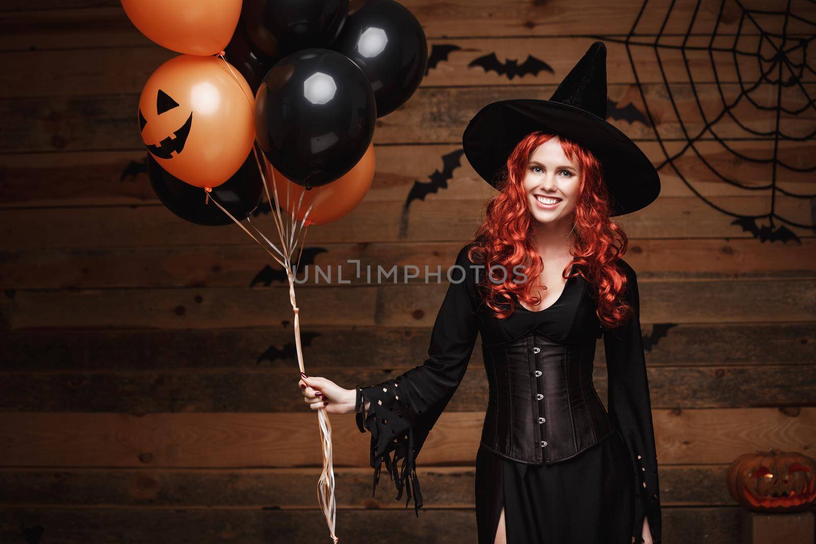 Halloween Witch Concept - Beautiful caucasian woman in witch costumes celebrating Halloween posing with posing with orange and black balloon over bats and spider web on Wooden studio background. by Benzoix