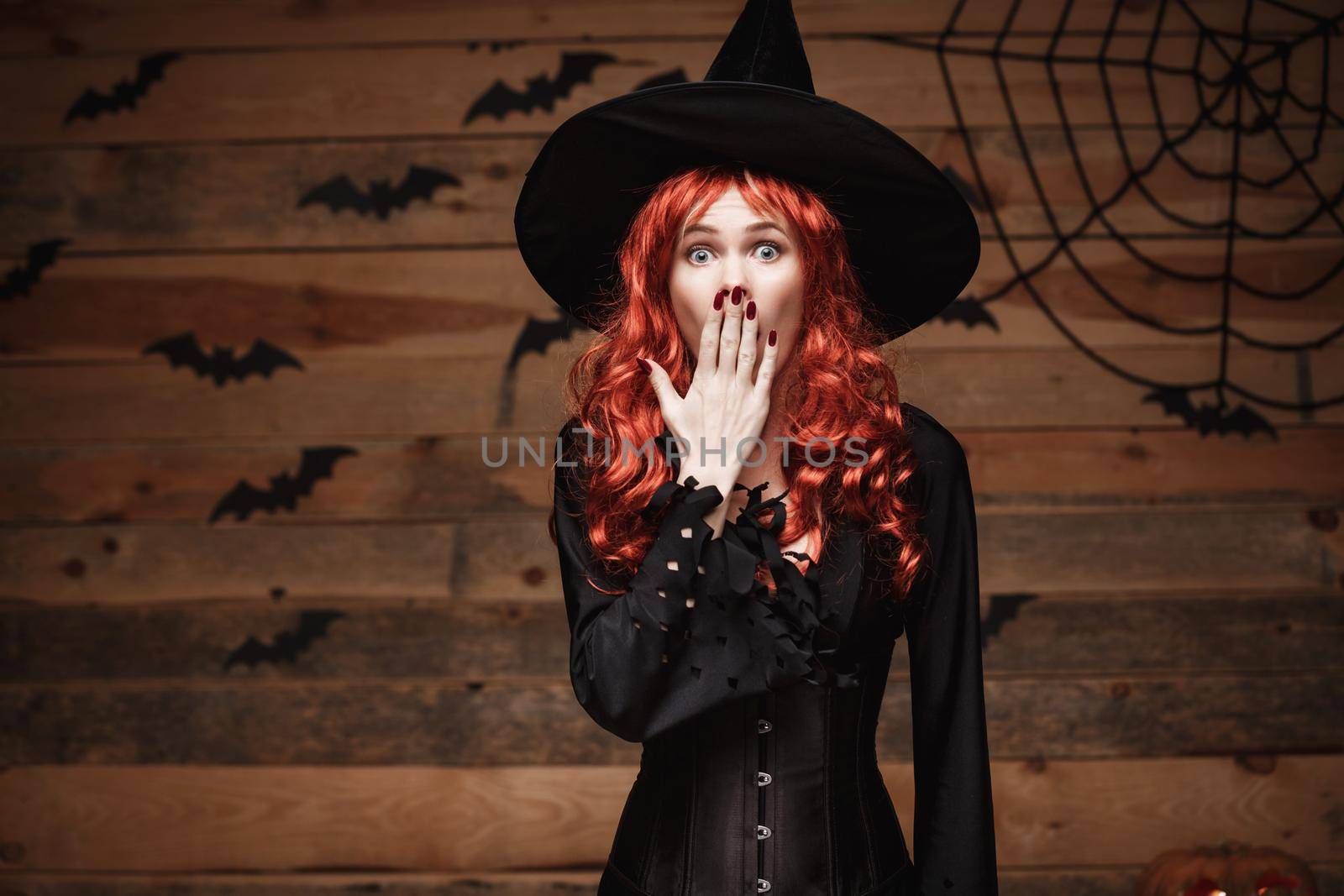 Halloween witch concept - Happy Halloween red hair Witch holding posing with shocked face over old wooden studio background. by Benzoix