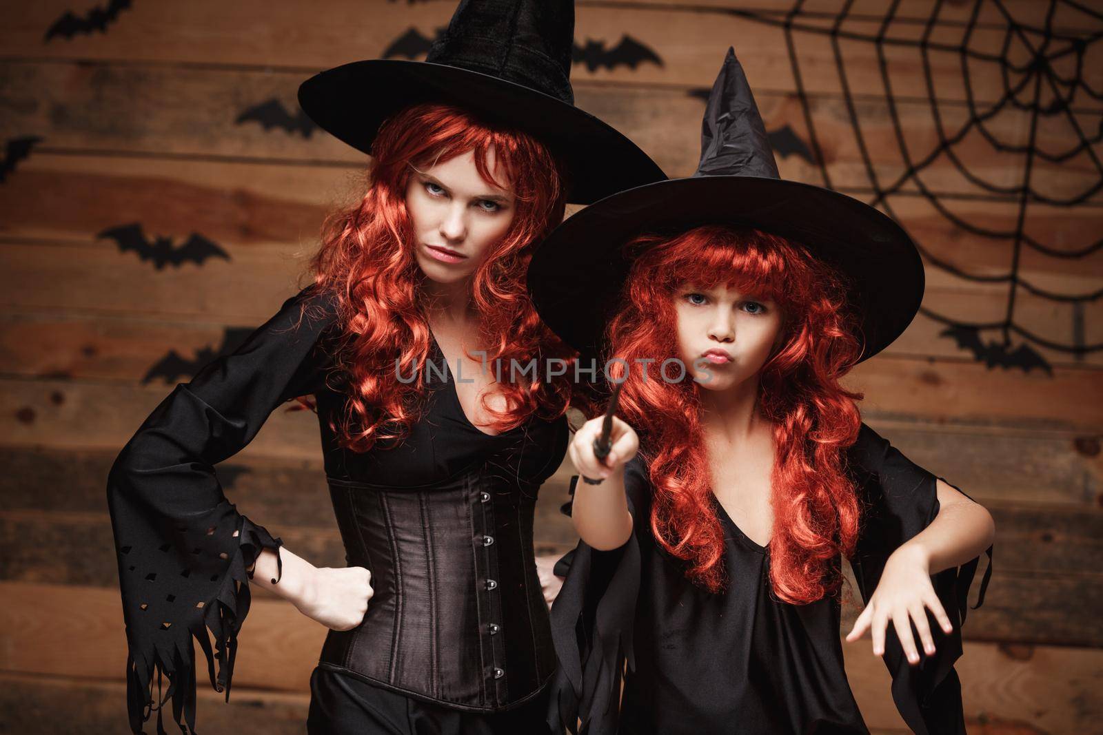 Beautiful caucasian mother and her daughter with long red hair in witch costumes and magic wand celebrating Halloween posing with over bats and spider web on Wooden studio background. by Benzoix