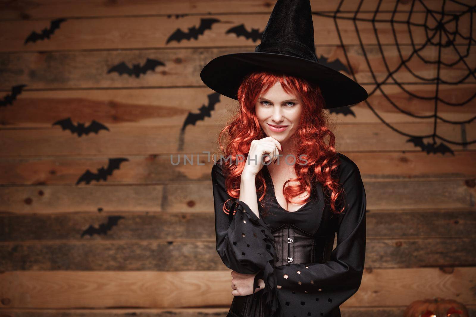 Halloween witch concept - Happy Halloween red hair Witch holding hand on her chin posing over old wooden studio background