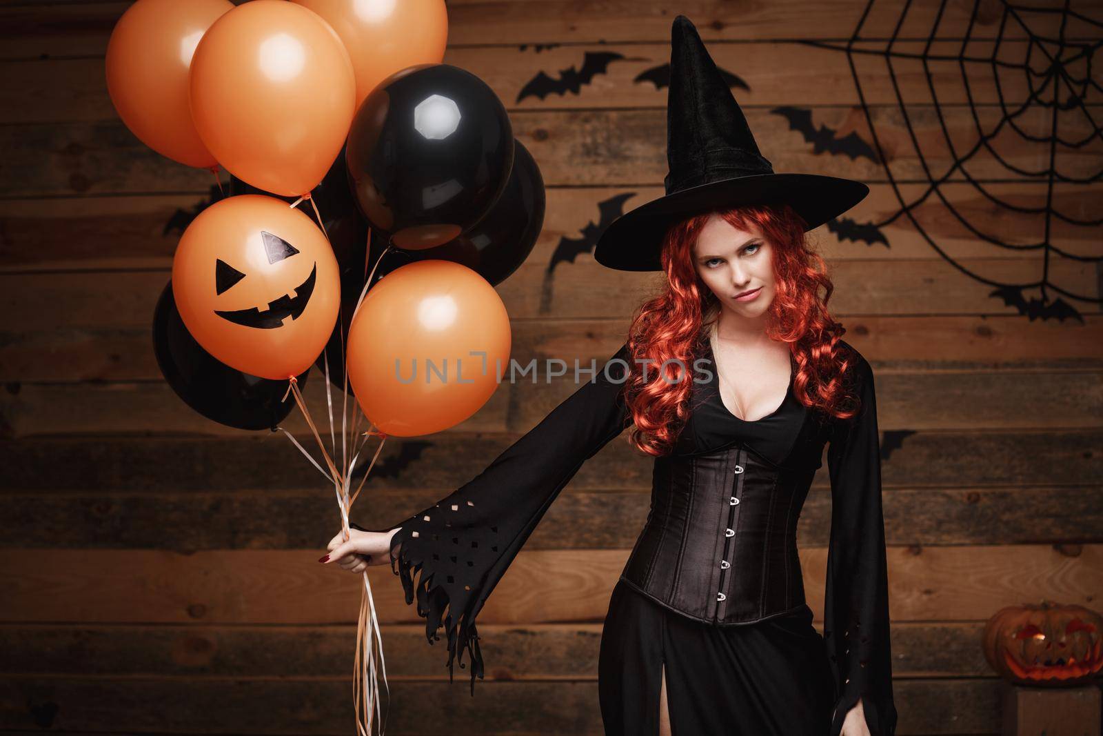 Beautiful caucasian woman in witch costumes celebrating Halloween posing with posing with orange and black balloon over bats and spider web on Wooden studio background