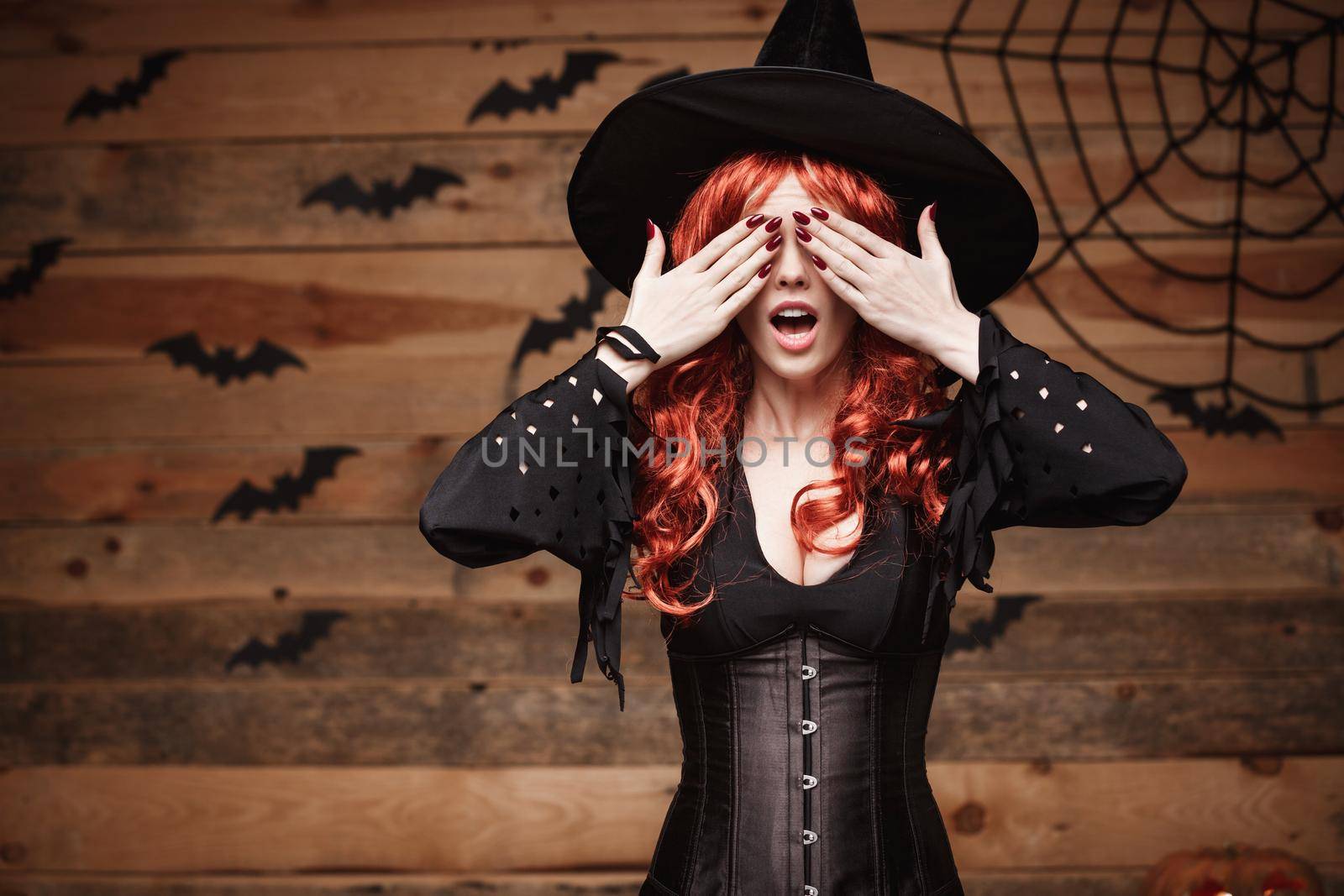 Halloween witch concept - Happy Halloween red hair Witch holding hands closing eyes posing over old wooden studio background. by Benzoix