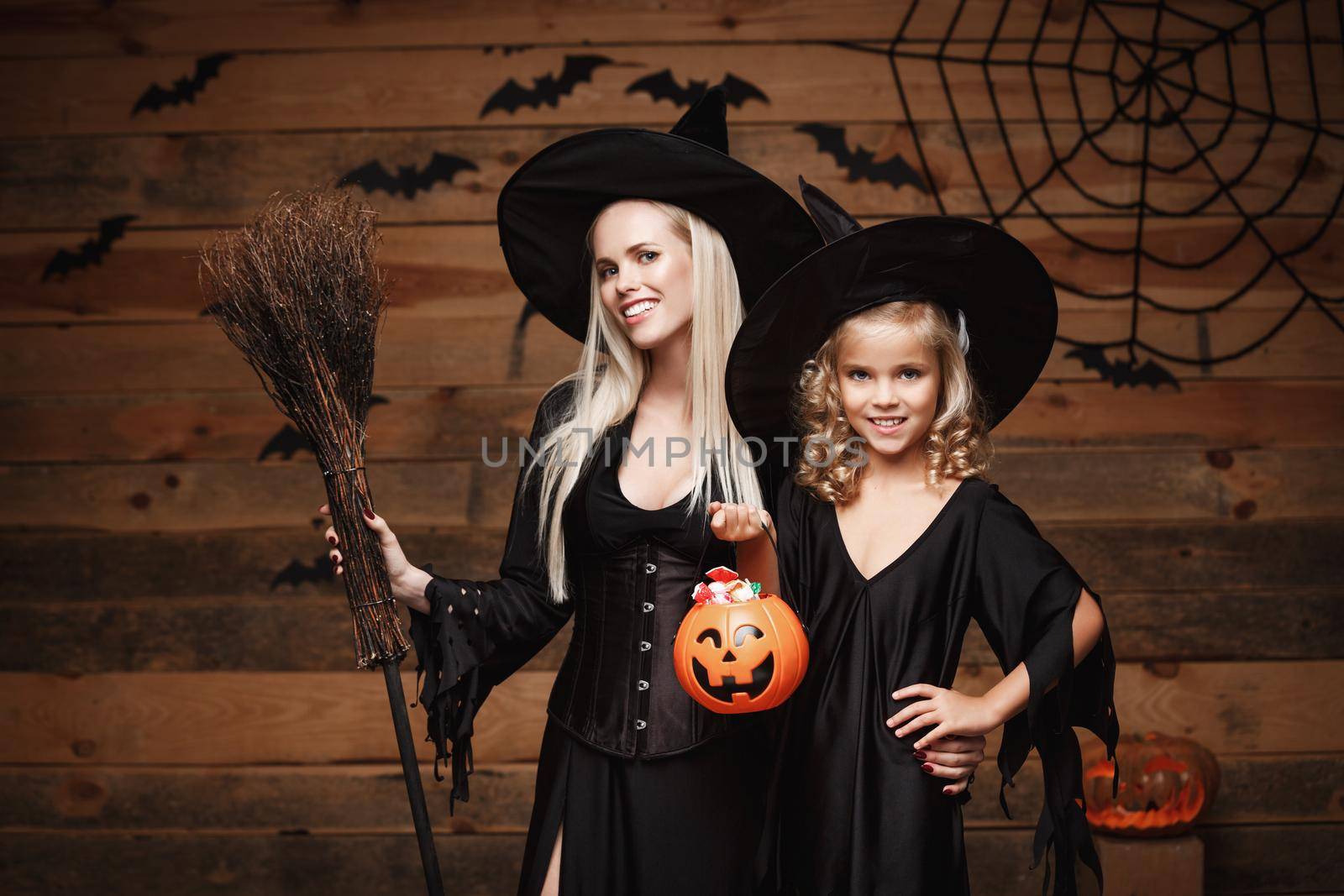 Halloween Concept: beautiful caucasian mother and her daughter in witch costumes celebrating Halloween with sharing Halloween candy and sweet over bats and spider web on Wooden studio background.