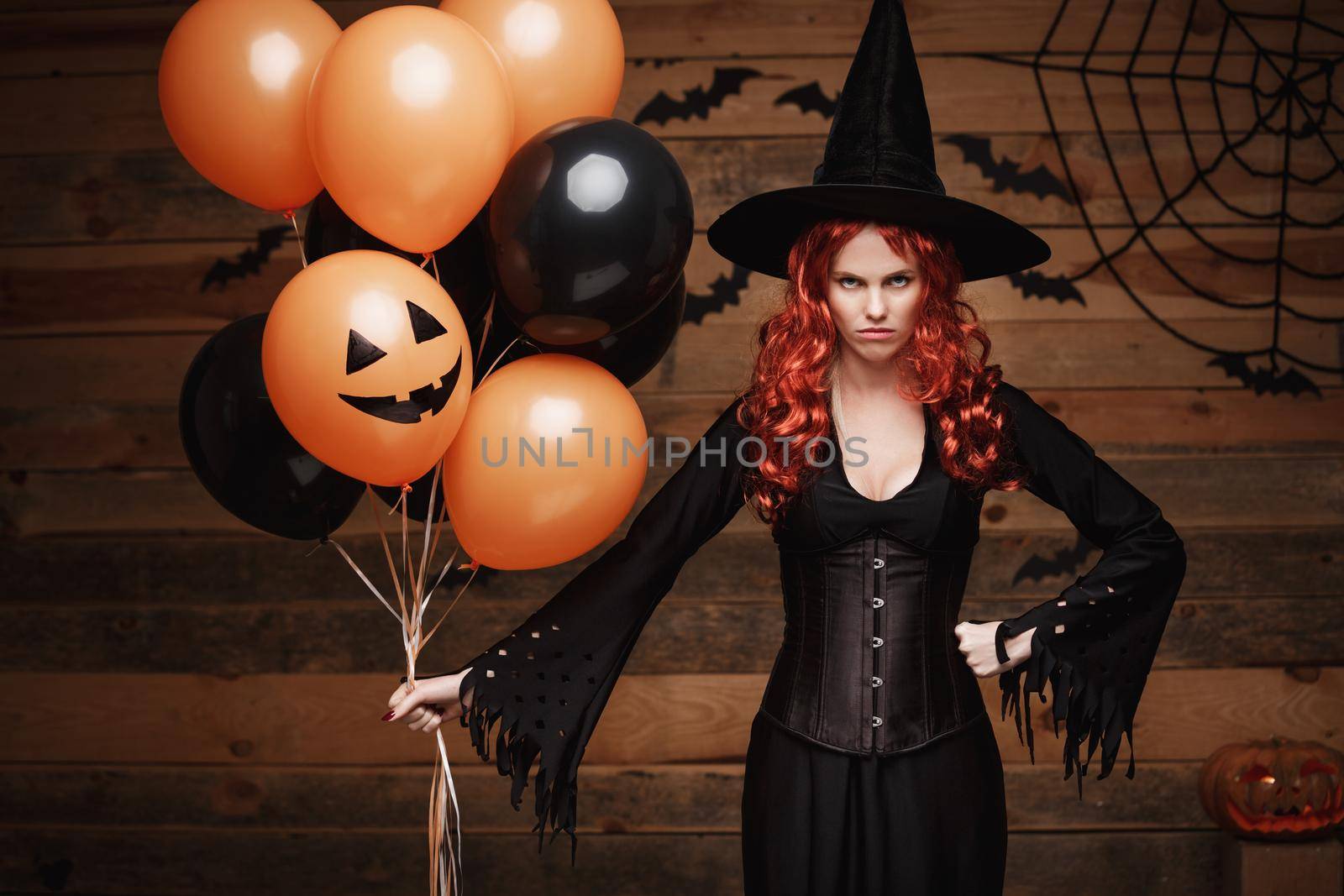 Beautiful caucasian woman in witch costumes celebrating Halloween posing with posing with orange and black balloon over bats and spider web on Wooden studio background