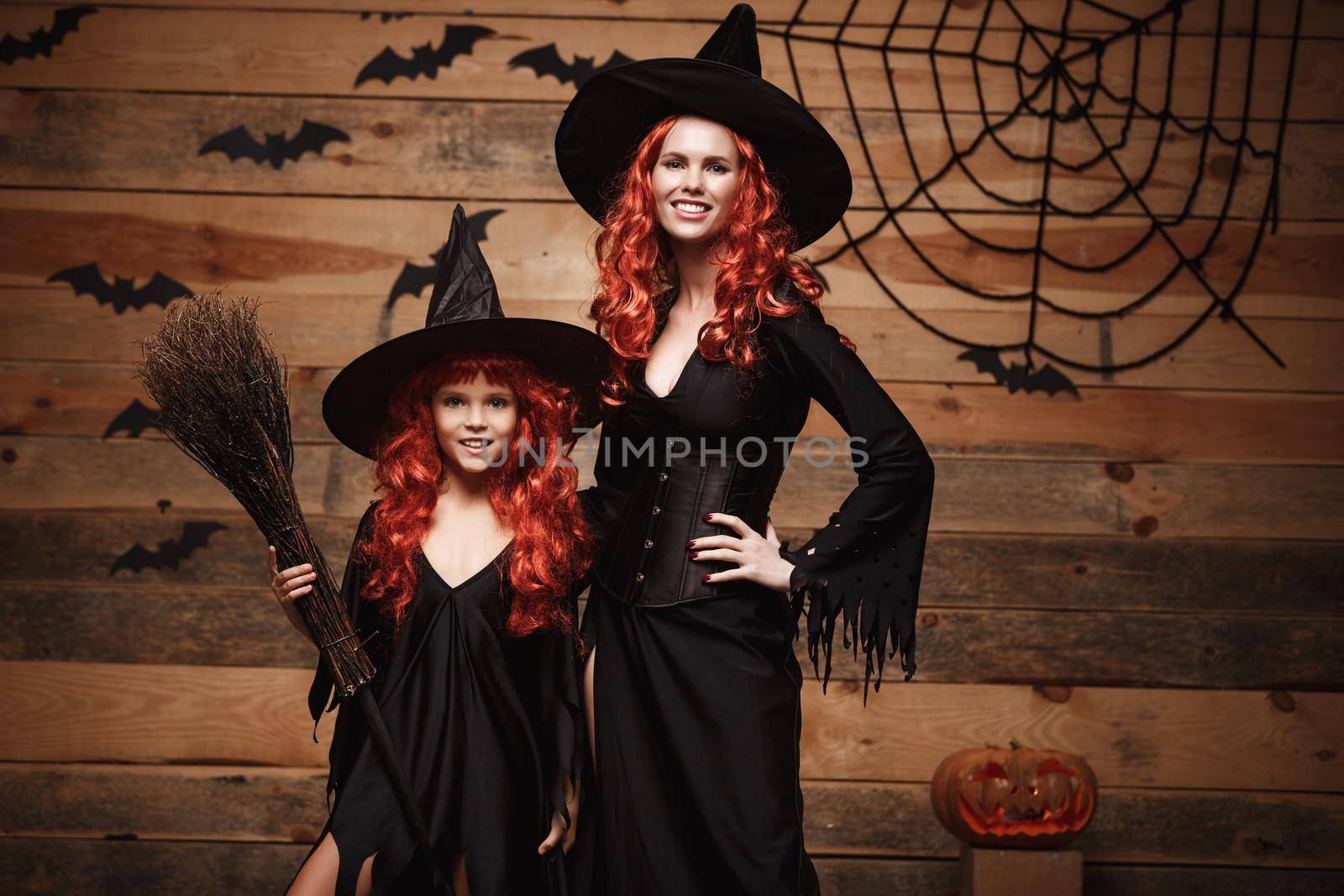 Halloween Concept - Beautiful caucasian mother and her daughter with long red hair in witch costumes celebrating Halloween posing with over bats and spider web on Wooden studio background. by Benzoix