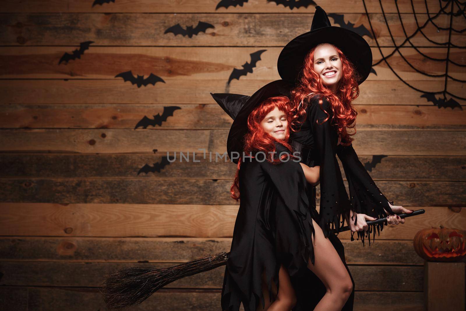 Halloween Concept - Beautiful caucasian mother and her daughter with long red hair in witch costumes flying with magic broomstick. by Benzoix