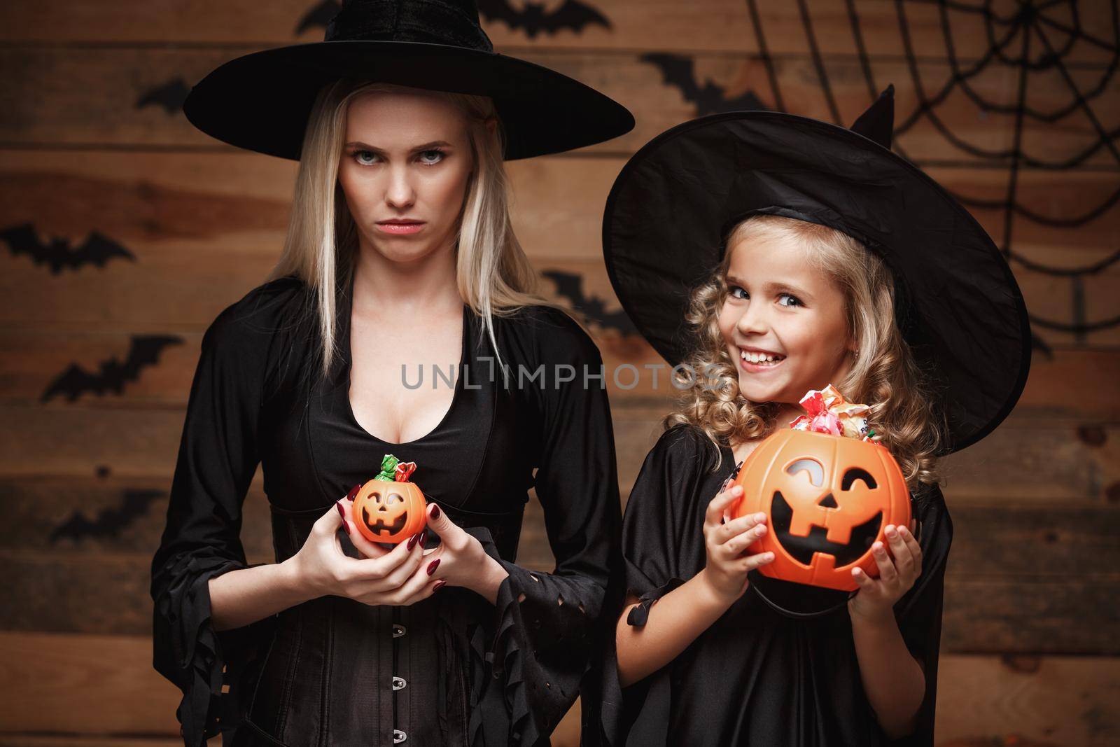 Halloween Concept: beautiful caucasian mother with disappointed feeling with happy little daugther enjoy with Halloween candy and sweet over bats and spider web on Wooden studio background