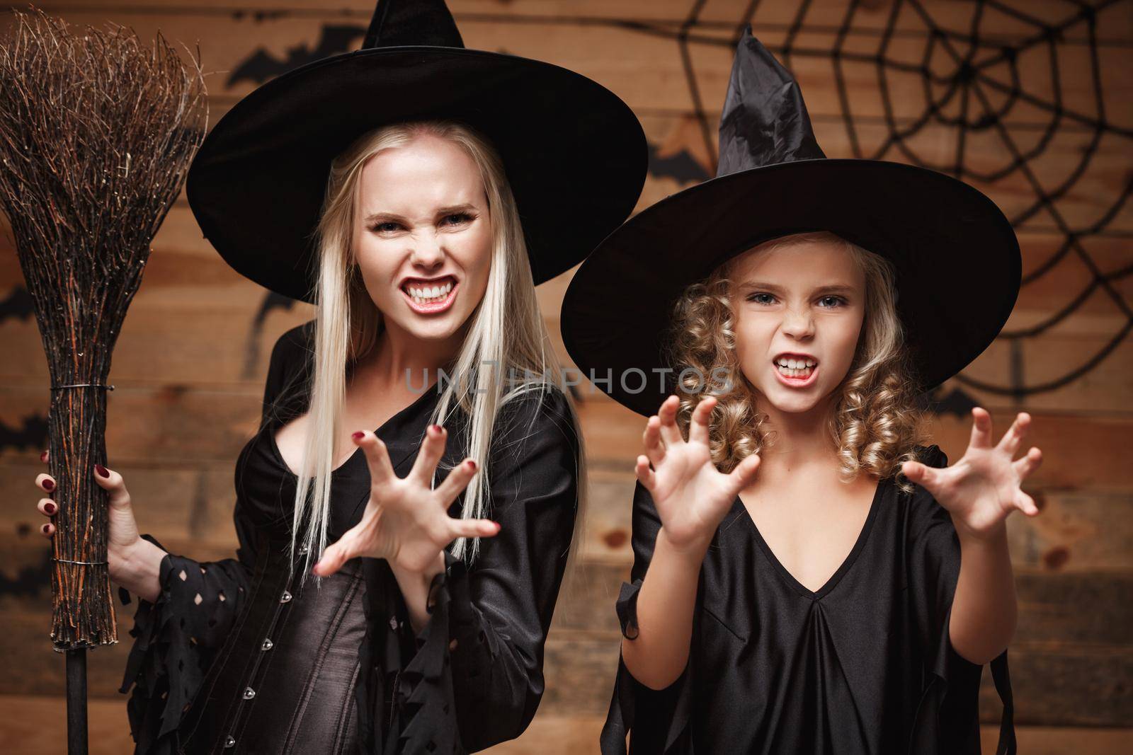 Halloween Concept - Closeup beautiful caucasian mother and her daughter in witch costumes celebrating Halloween posing with curved pumpkins over bats and spider web on Wooden studio background. by Benzoix