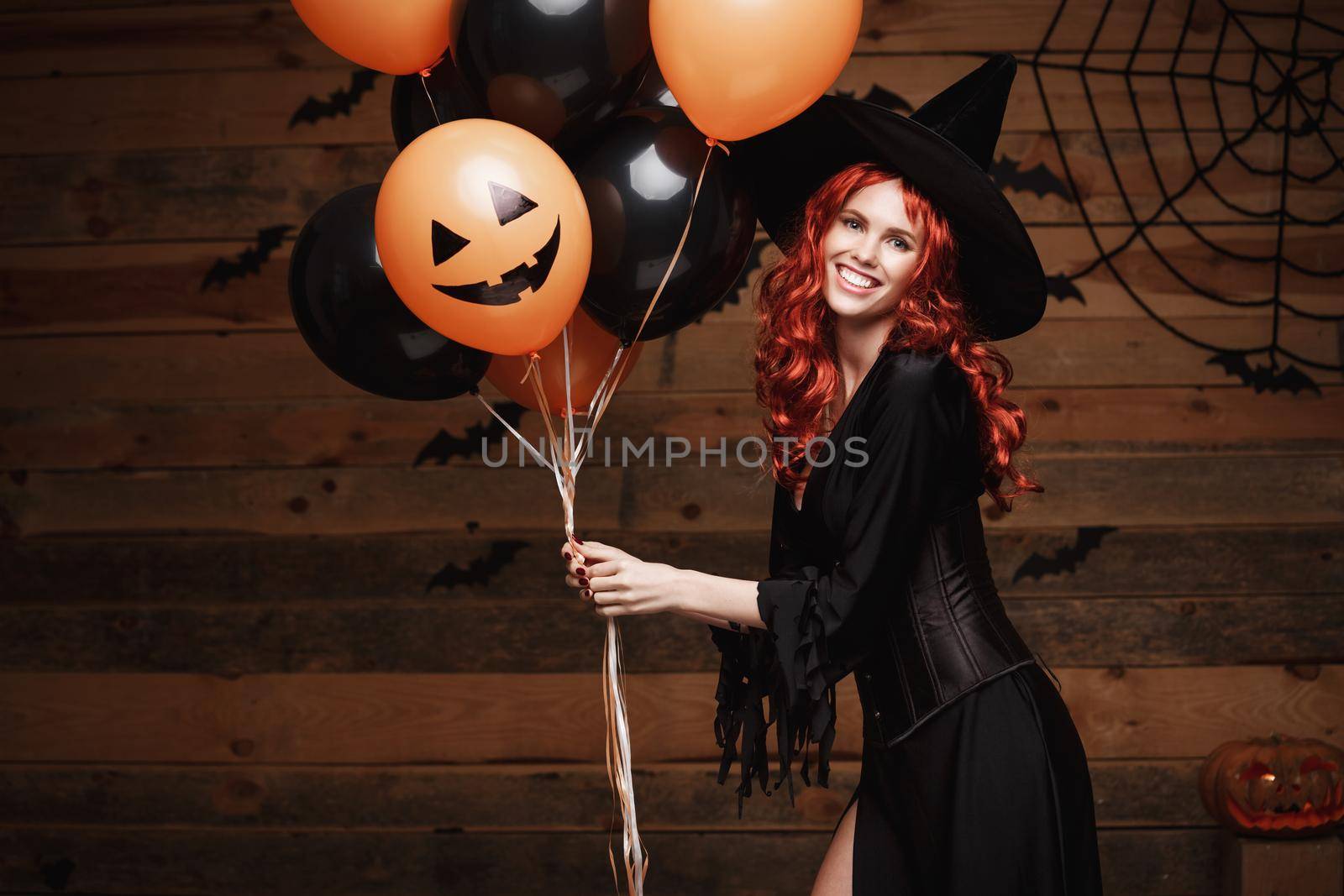 Halloween Witch Concept - Beautiful caucasian woman in witch costumes celebrating Halloween posing with posing with orange and black balloon over bats and spider web on Wooden studio background. by Benzoix