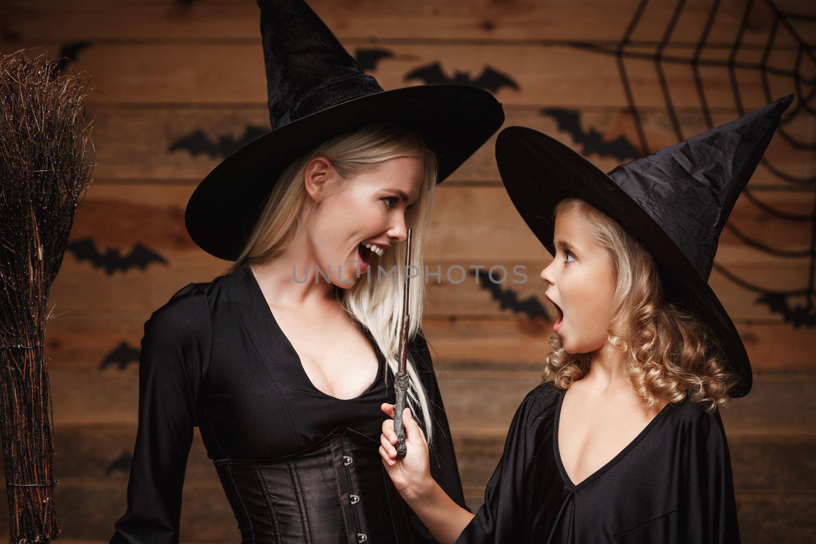 Halloween Concept - Closeup beautiful caucasian mother and her daughter in witch costumes celebrating Halloween posing with curved pumpkins over bats and spider web on Wooden studio background. by Benzoix