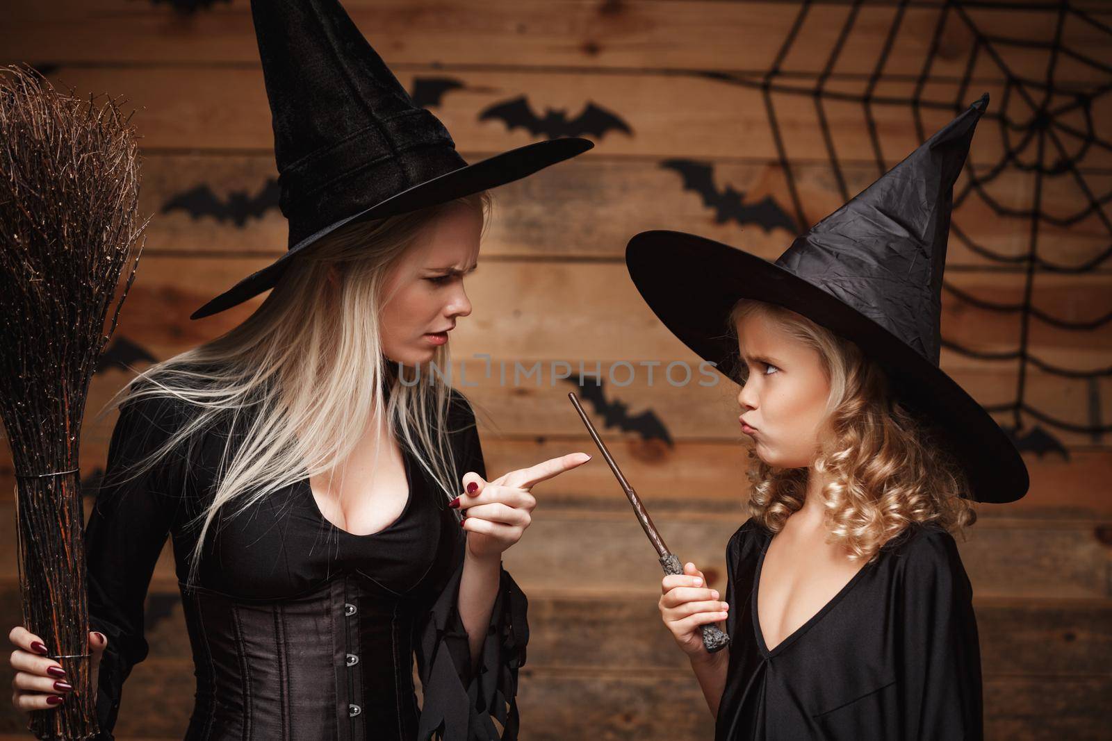 Halloween Concept - stressful witch mother teaching her daughter in witch costumes celebrating Halloween over bats and spider web on Wooden studio background