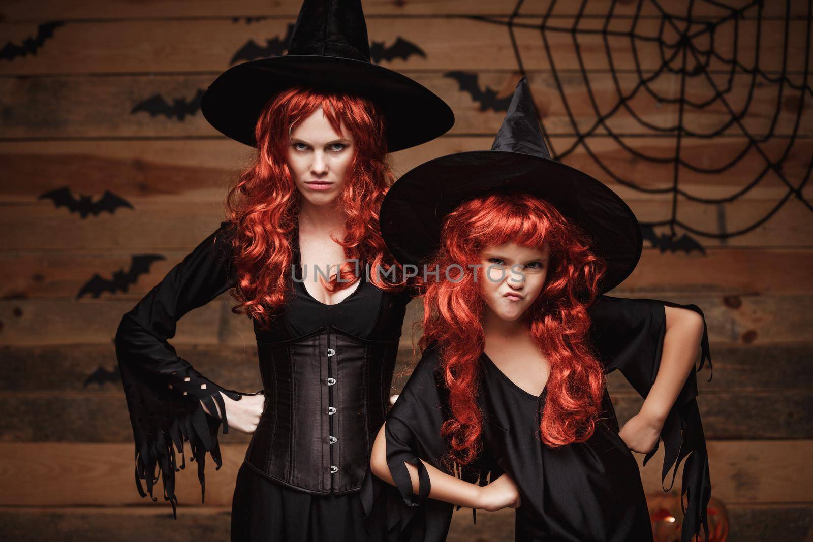 Halloween Concept - Beautiful caucasian mother and her daughter with long red hair in witch costumes with angry fussy facial expression. by Benzoix