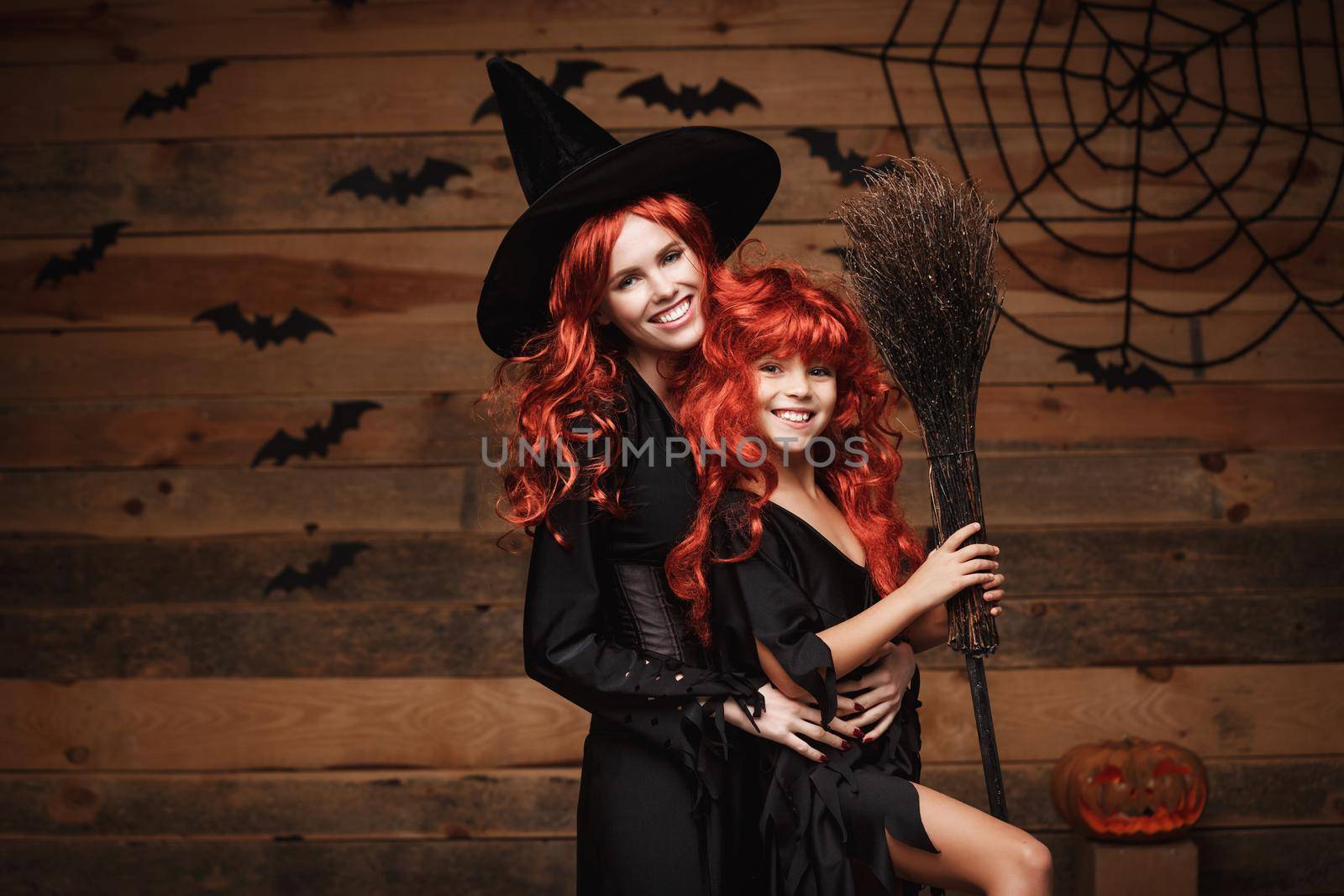 Beautiful caucasian mother and her daughter with long red hair in witch costumes celebrating Halloween posing with over bats and spider web on Wooden studio background. by Benzoix