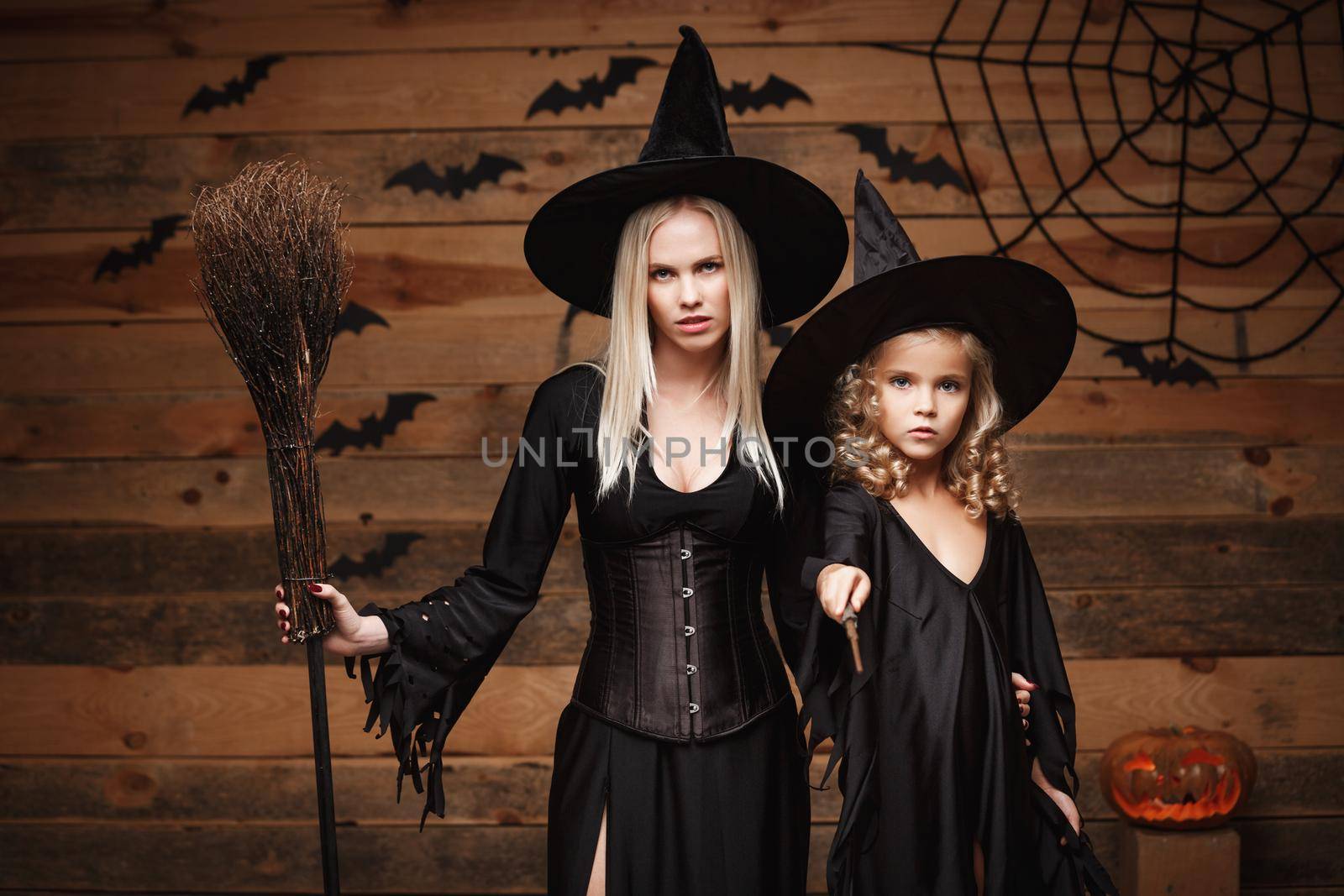 Halloween Concept - cheerful mother and her daughter in witch costumes celebrating Halloween posing with curved pumpkins over bats and spider web on Wooden studio background