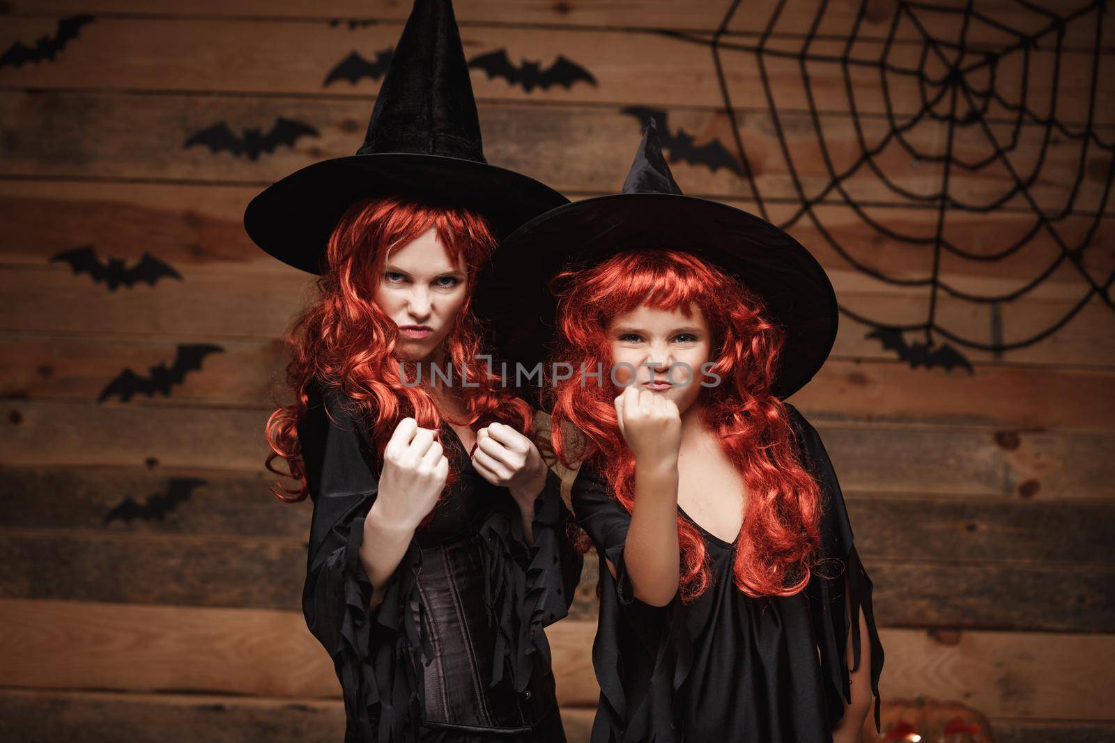 Halloween Concept - Beautiful caucasian mother and her daughter with long red hair in witch costumes witch cheerful fighting facial expression. by Benzoix