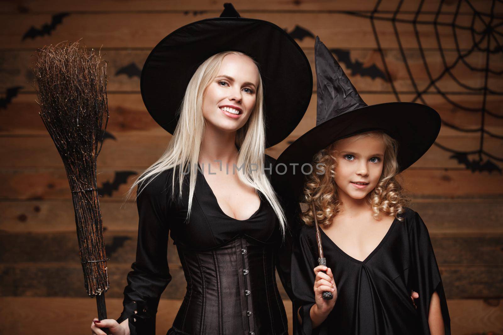 Halloween Concept - Closeup beautiful caucasian mother and her daughter in witch costumes celebrating Halloween posing with curved pumpkins over bats and spider web on Wooden studio background. by Benzoix