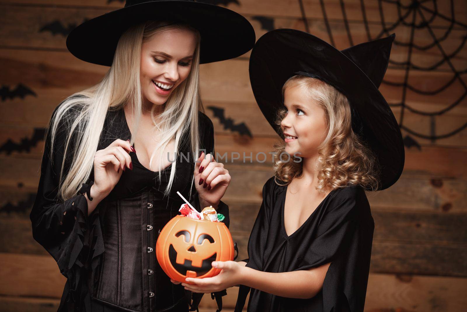 Halloween Concept: beautiful caucasian mother and her daughter in witch costumes celebrating Halloween with sharing Halloween candy and sweet over bats and spider web on Wooden studio background