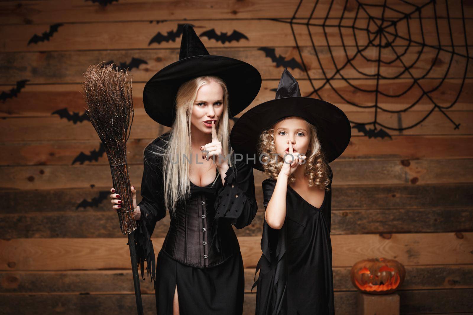 Halloween Concept - cheerful mother and her daughter in witch costumes celebrating Halloween doing silence gesture posing with curved pumpkins over bats and spider web on Wooden studio background. by Benzoix