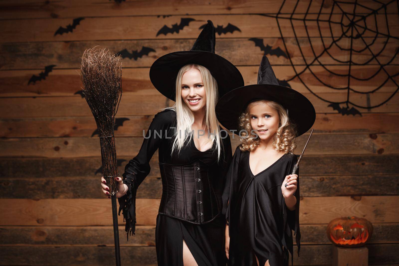 Halloween Concept - cheerful mother and her daughter in witch costumes celebrating Halloween posing with curved pumpkins over bats and spider web on Wooden studio background