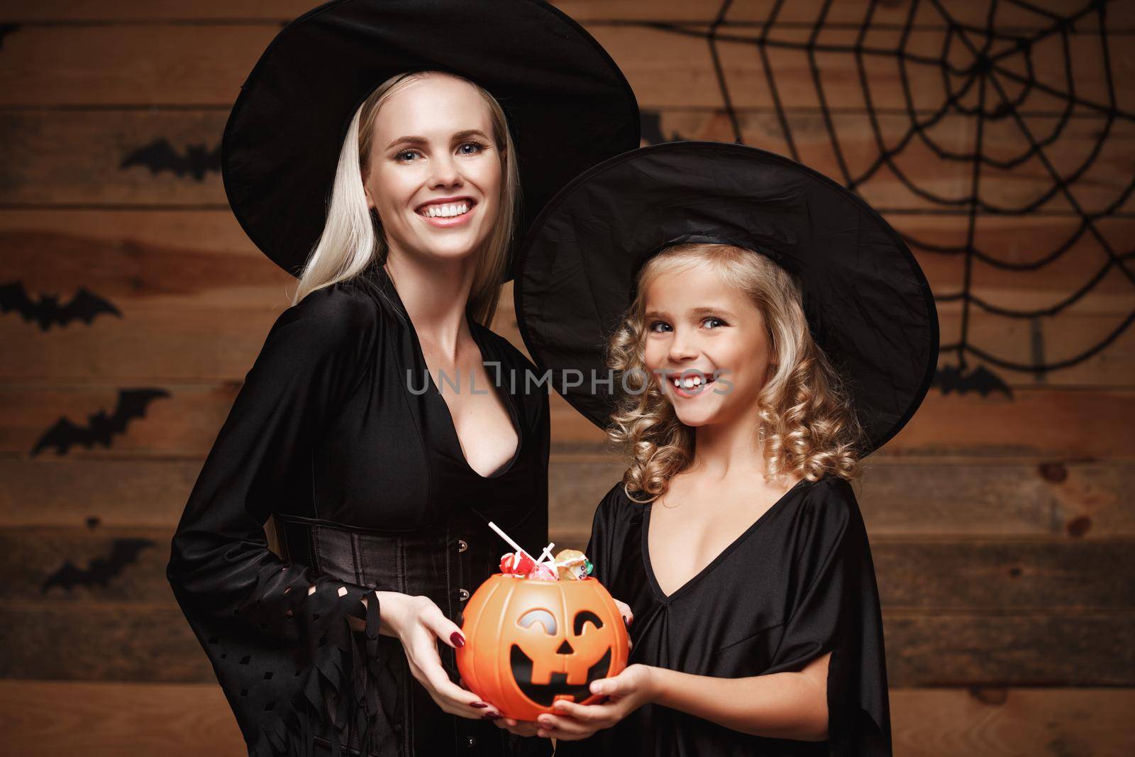 Halloween Concept: beautiful caucasian mother and her daughter in witch costumes celebrating Halloween with sharing Halloween candy and sweet over bats and spider web on Wooden studio background