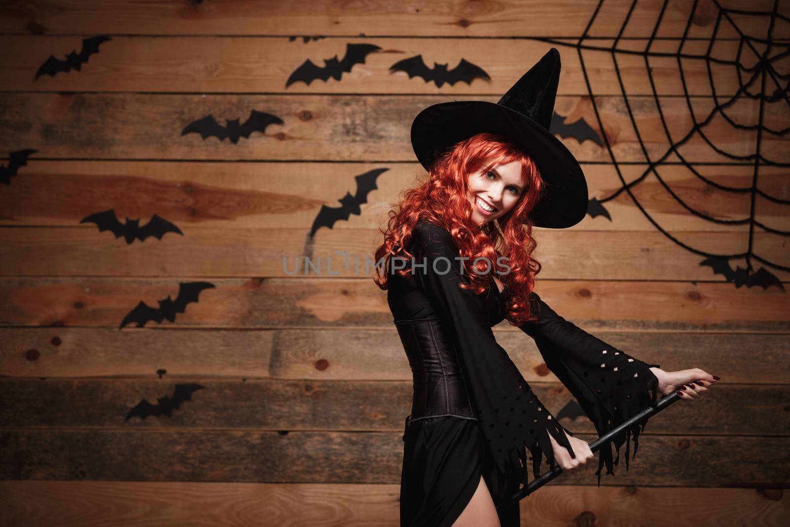 Halloween witch concept - Happy Halloween red hair Witch holding magic broomstick flying gesture over old wooden studio background. by Benzoix