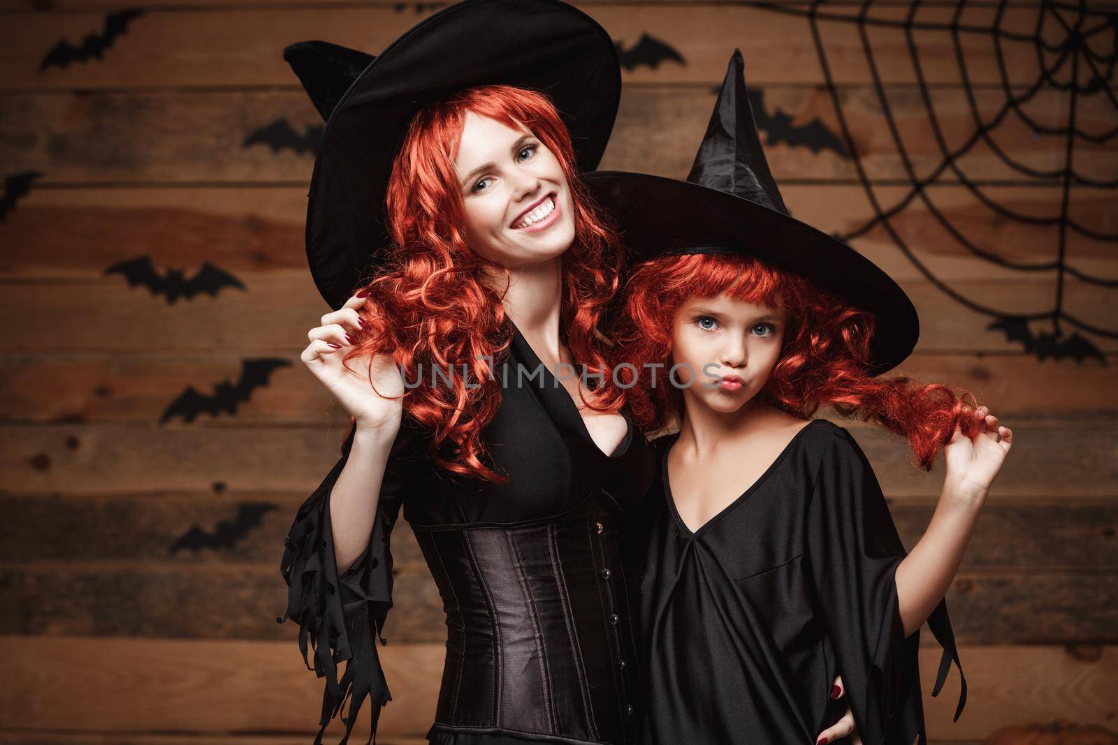 Halloween Concept - Beautiful caucasian mother and her daughter with long red hair in witch costumes celebrating Halloween posing with over bats and spider web on Wooden studio background. by Benzoix