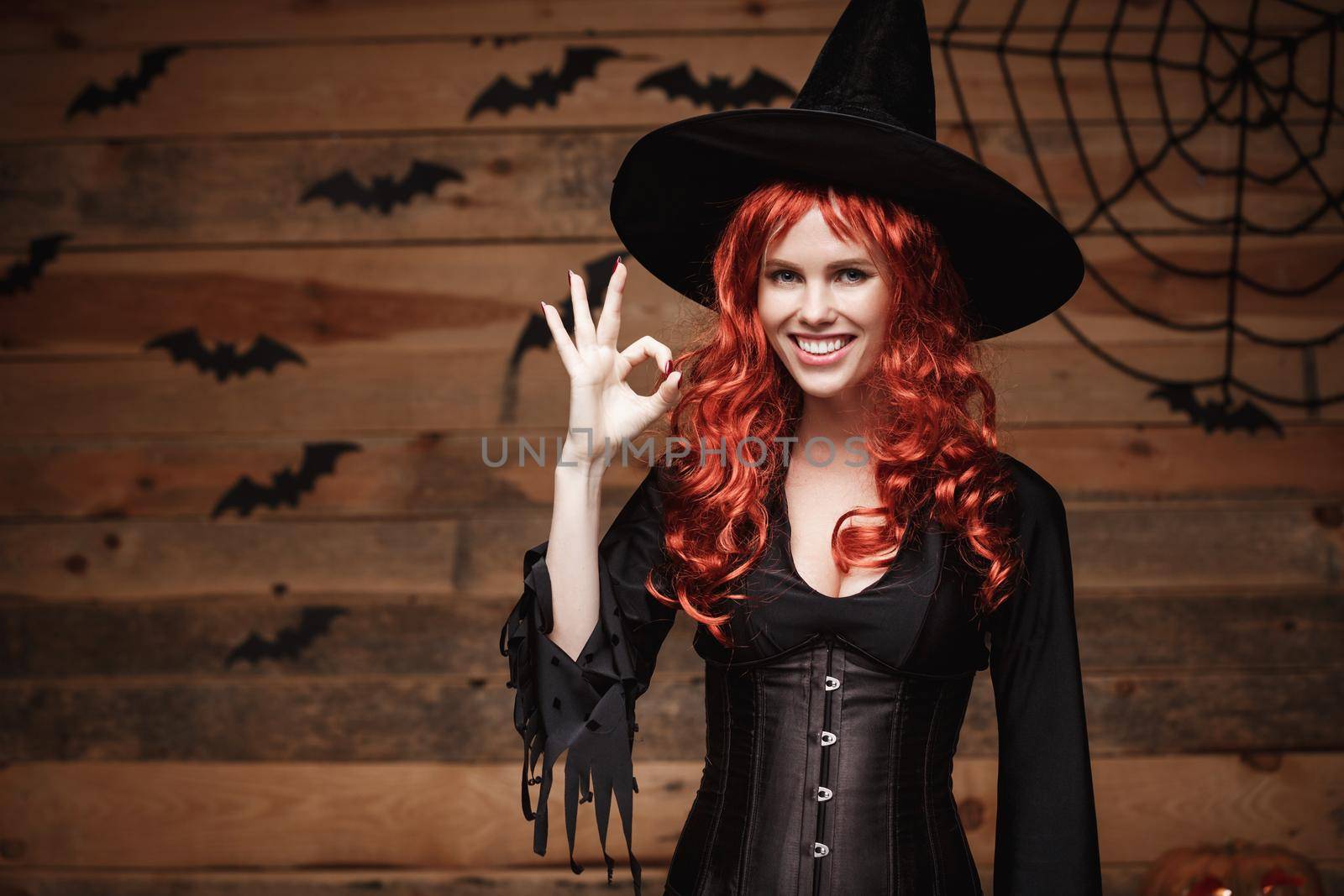 Halloween witch concept - Happy Halloween red hair Witch holding ok sign with fingers posing over old wooden studio background