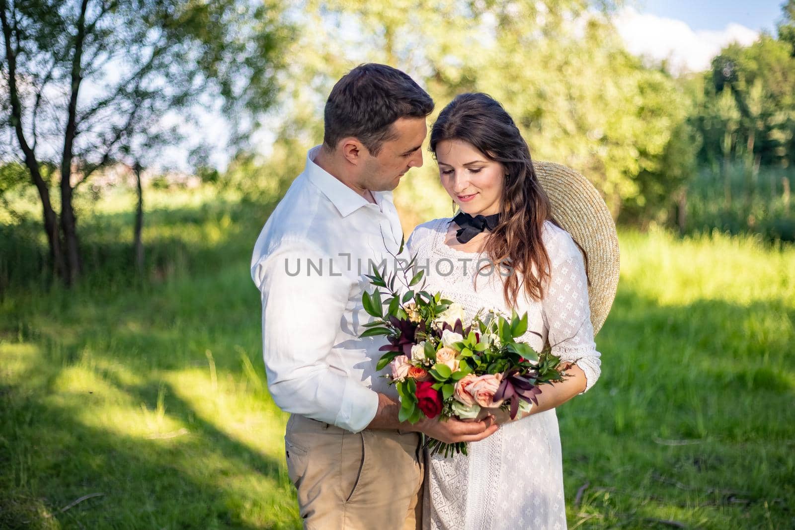 Happy young people in wedding dresses stand in the meadow and look at each other. portrait of a bride who looks at a beautiful wedding bouquet and her husband looks at his wife by Anyatachka