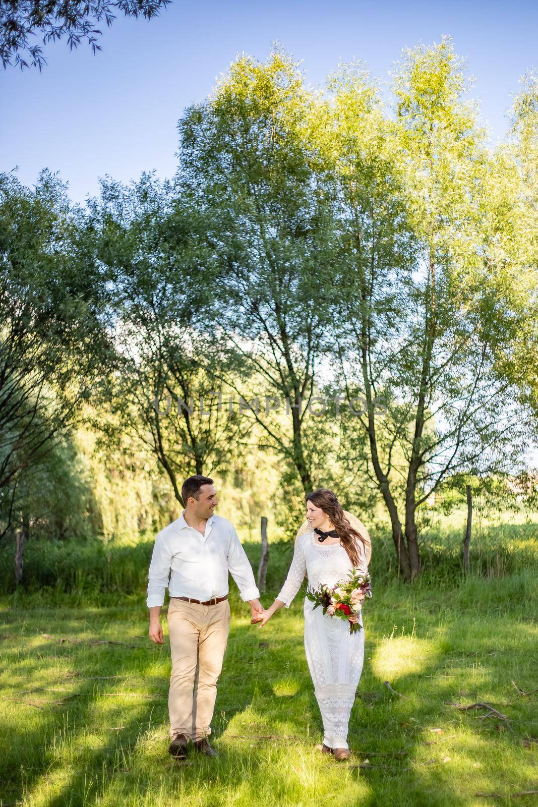 Happy young people in wedding attire walk in the meadow and look at each other by Anyatachka