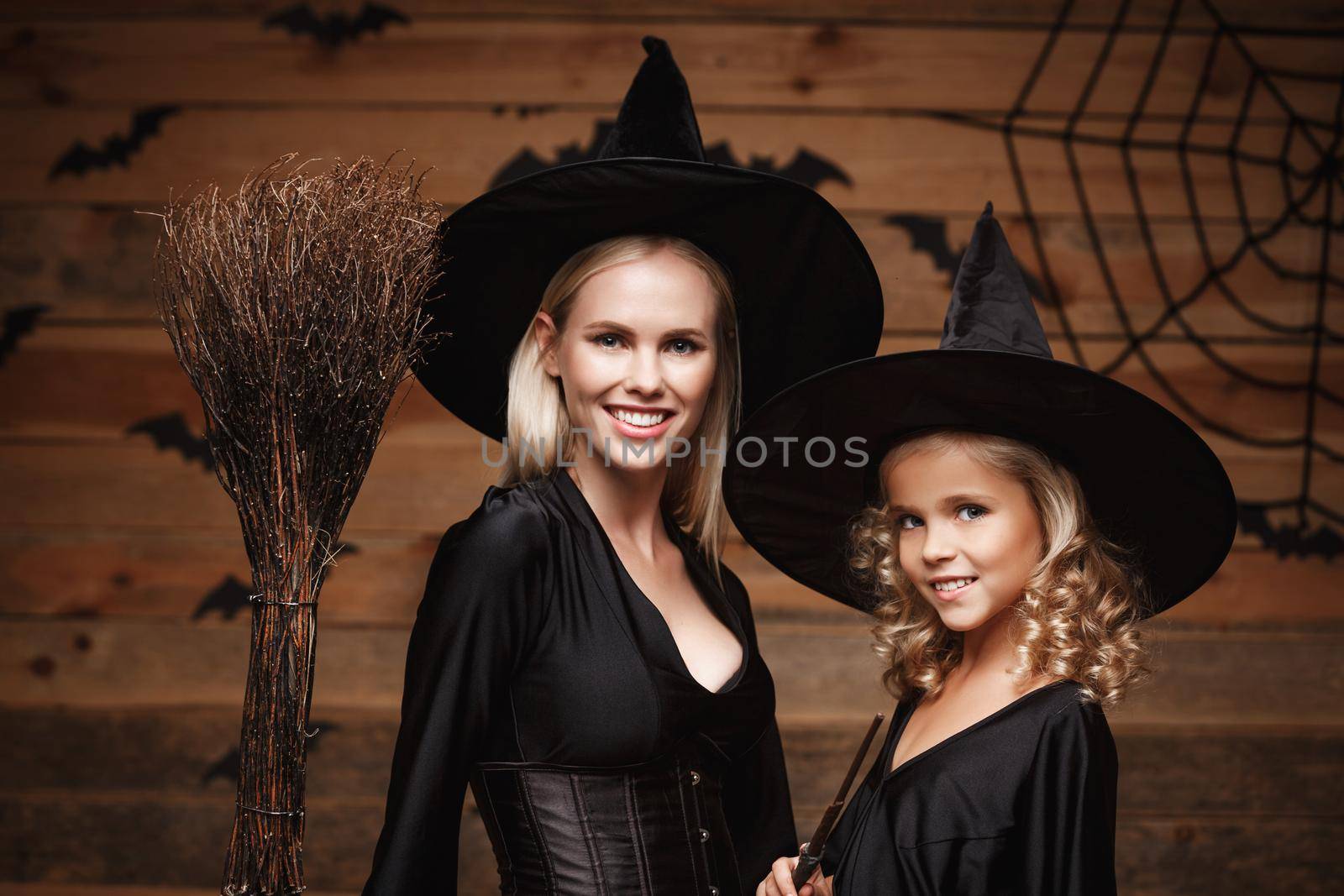 Halloween Concept - Closeup beautiful caucasian mother and her daughter in witch costumes celebrating Halloween posing with curved pumpkins over bats and spider web on Wooden studio background. by Benzoix