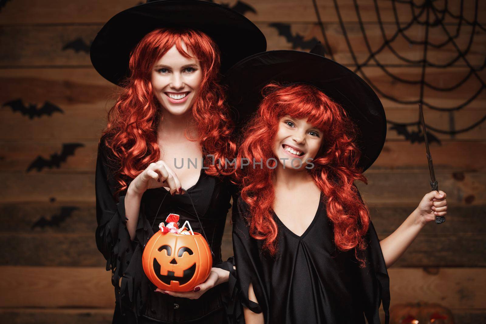 Halloween Concept - Beautiful caucasian mother and her daughter with long red hair in witch costumes with Halloween candy and magic wand. by Benzoix