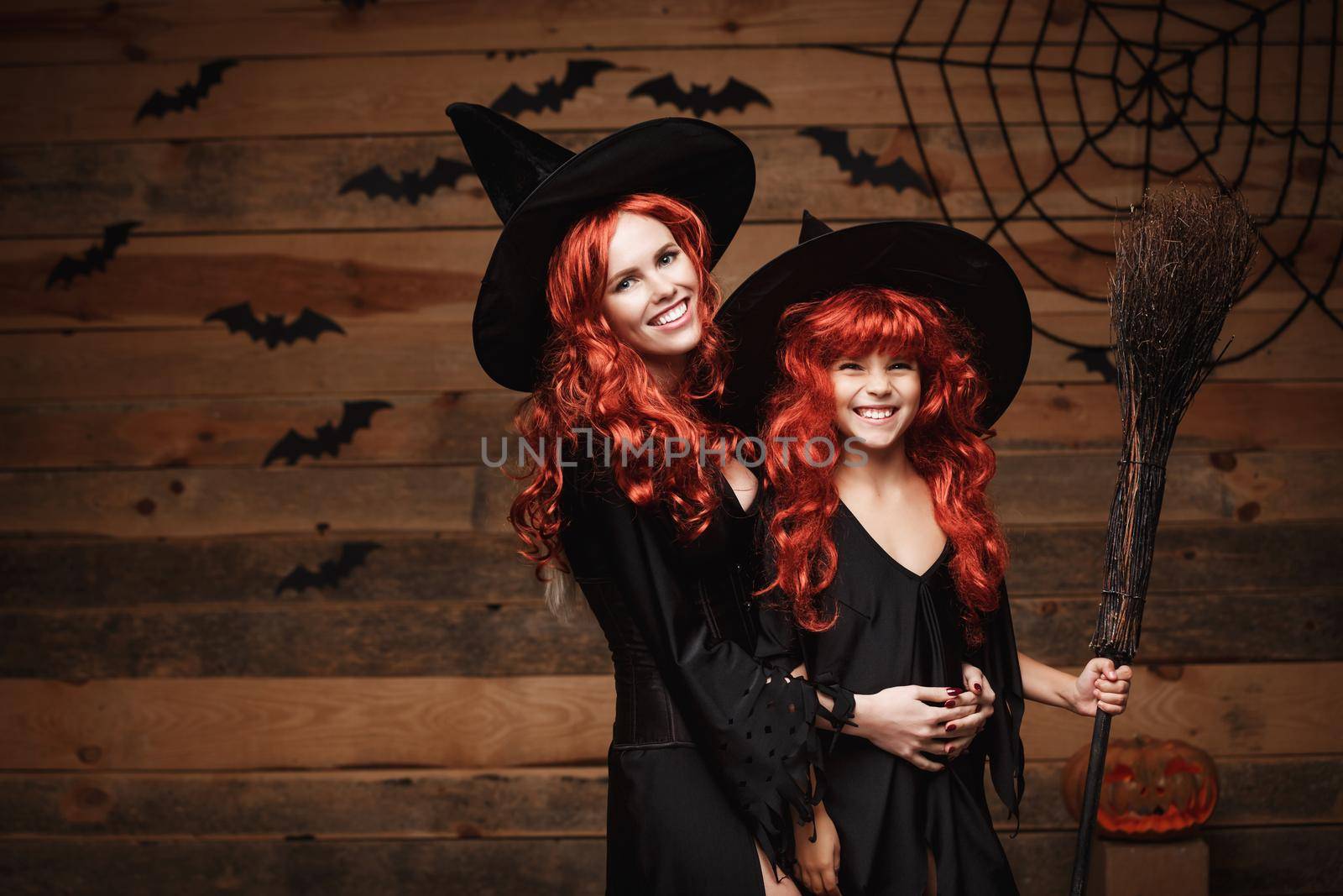 Beautiful caucasian mother and her daughter with long red hair in witch costumes celebrating Halloween posing with over bats and spider web on Wooden studio background. by Benzoix