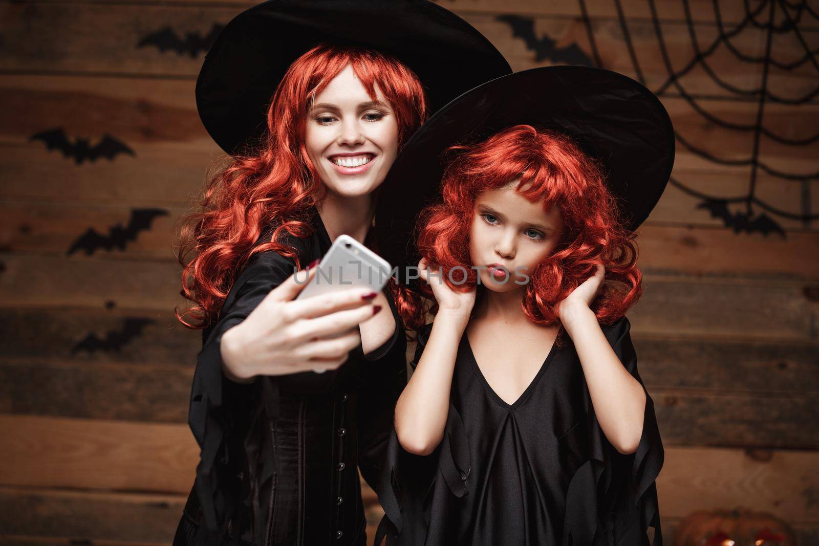 Halloween Concept - Beautiful caucasian mother and her daughter with long red hair in witch costumes taking a selfie with smartphone ready for celebrating Halloween. by Benzoix