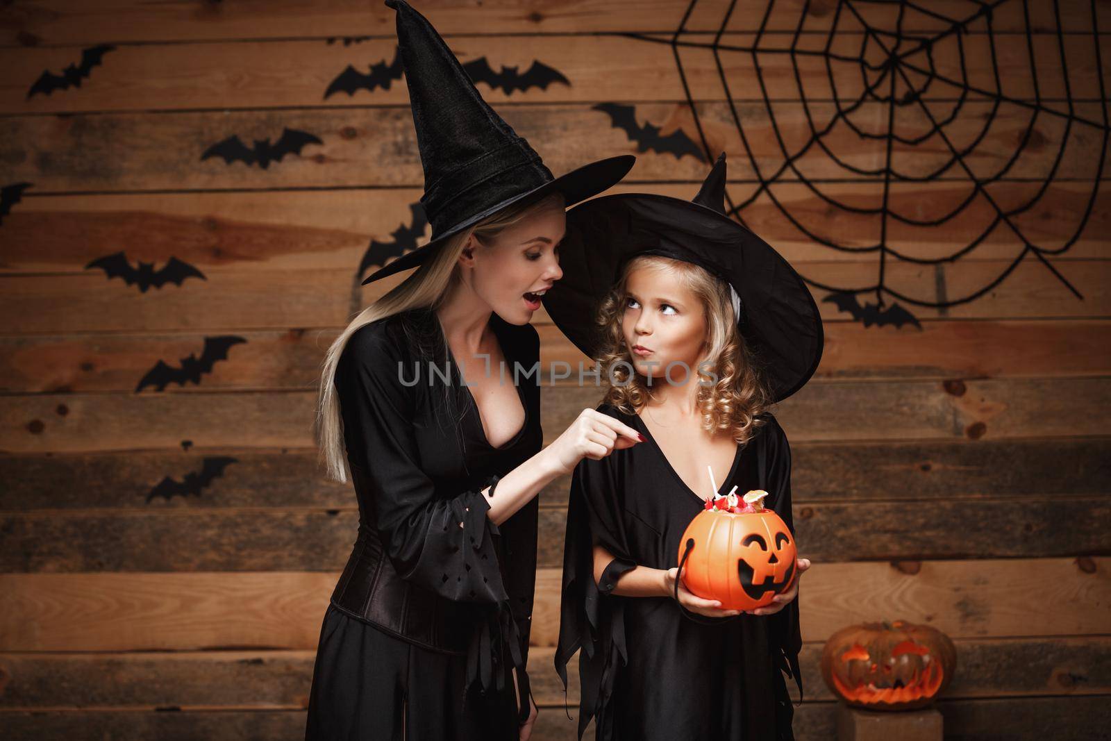 Halloween Concept - beautiful caucasian mother and her daughter in witch costumes celebrating Halloween with sharing Halloween candy and sweet over bats and spider web on Wooden studio background. by Benzoix