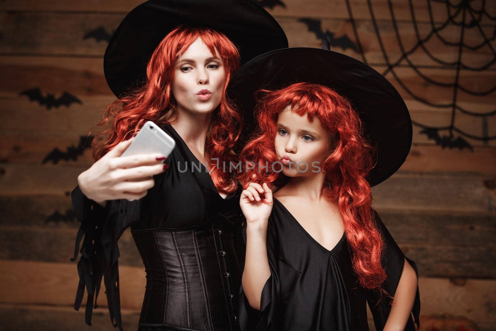 Halloween Concept - Beautiful caucasian mother and her daughter with long red hair in witch costumes taking a selfie with smartphone ready for celebrating Halloween. by Benzoix