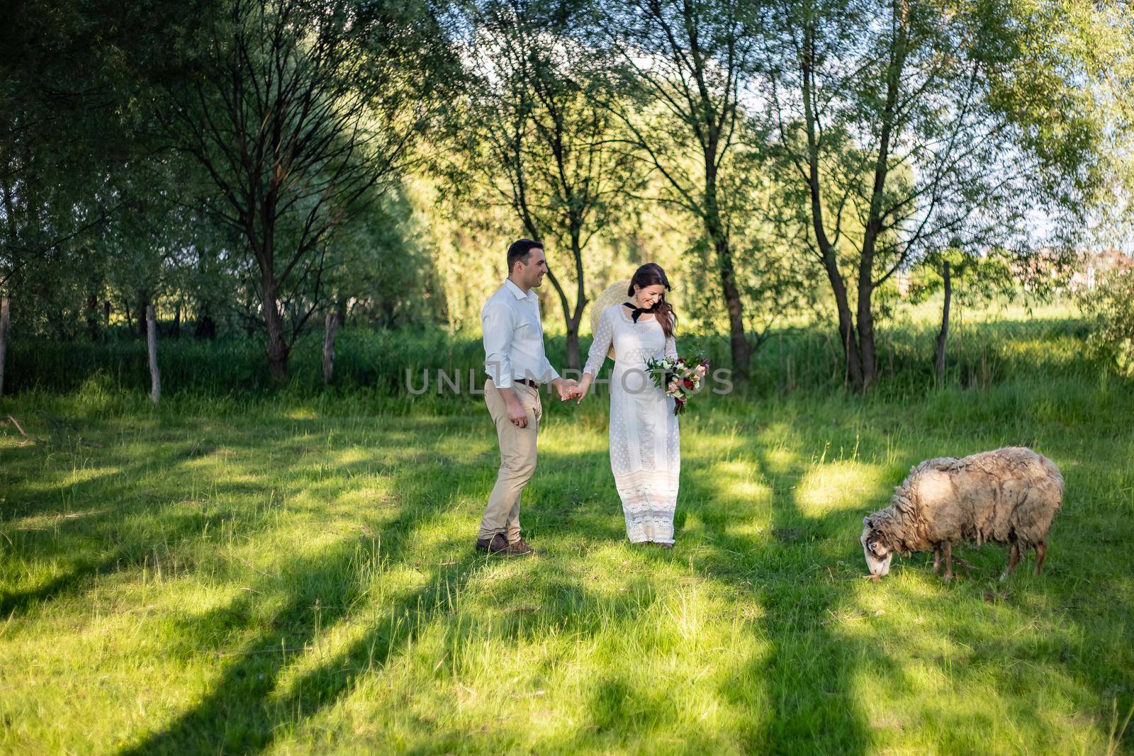 Happy young people in wedding dresses stand in the meadow and look at the lamb by Anyatachka