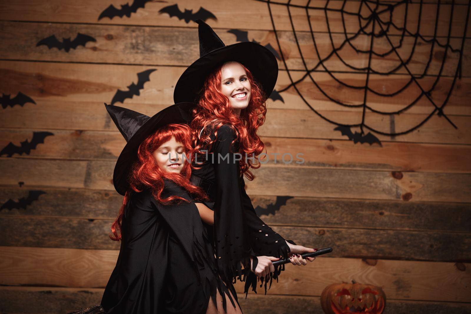 Halloween Concept - Beautiful caucasian mother and her daughter with long red hair in witch costumes flying with magic broomstick. by Benzoix