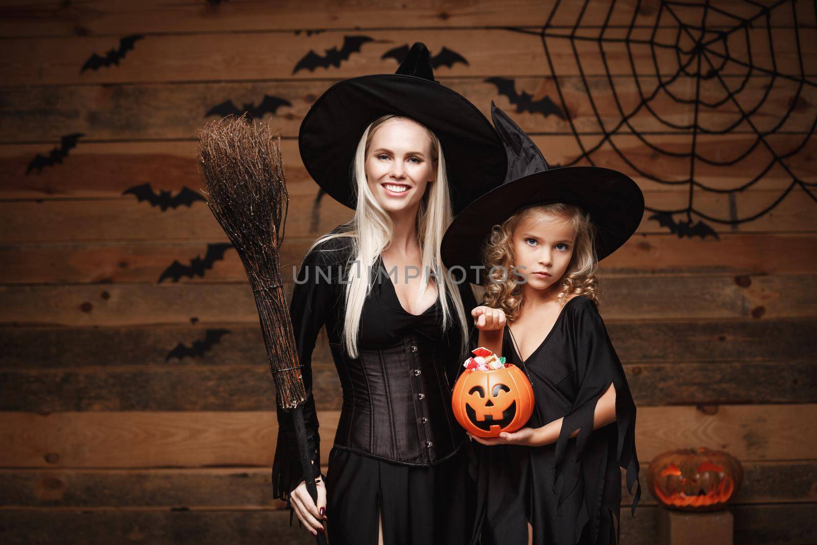 Halloween Concept: beautiful caucasian mother and her daughter in witch costumes celebrating Halloween with sharing Halloween candy and sweet over bats and spider web on Wooden studio background.