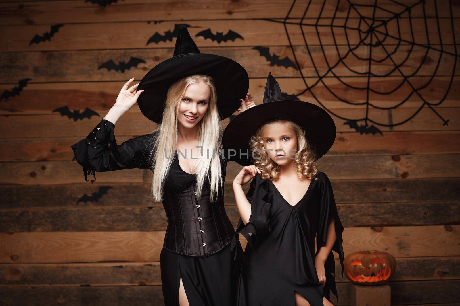 Halloween Concept - cheerful mother and her daughter in witch costumes celebrating Halloween posing with curved pumpkins over bats and spider web on Wooden studio background. by Benzoix