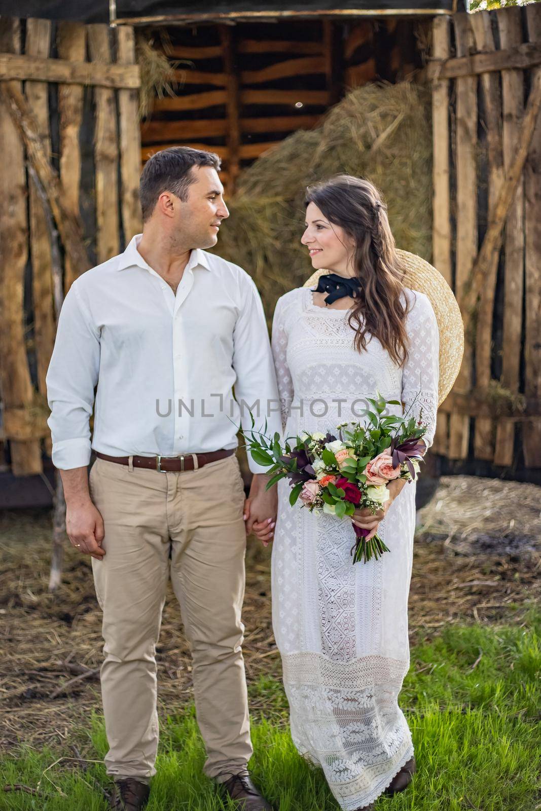 Stylishly dressed handsome groom with a beard in black sunglasses hugs wife. Bride in a dress with a bouquet puts a hat on and laughs. Rustic wedding in the style of boho at the ranch.
