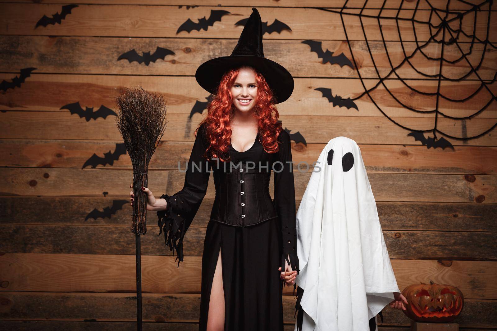 Halloween Concept: Witch mother and little white ghost doing trick or treat celebrating Halloween posing with curved pumpkins over bats and spider web on Wooden studio background