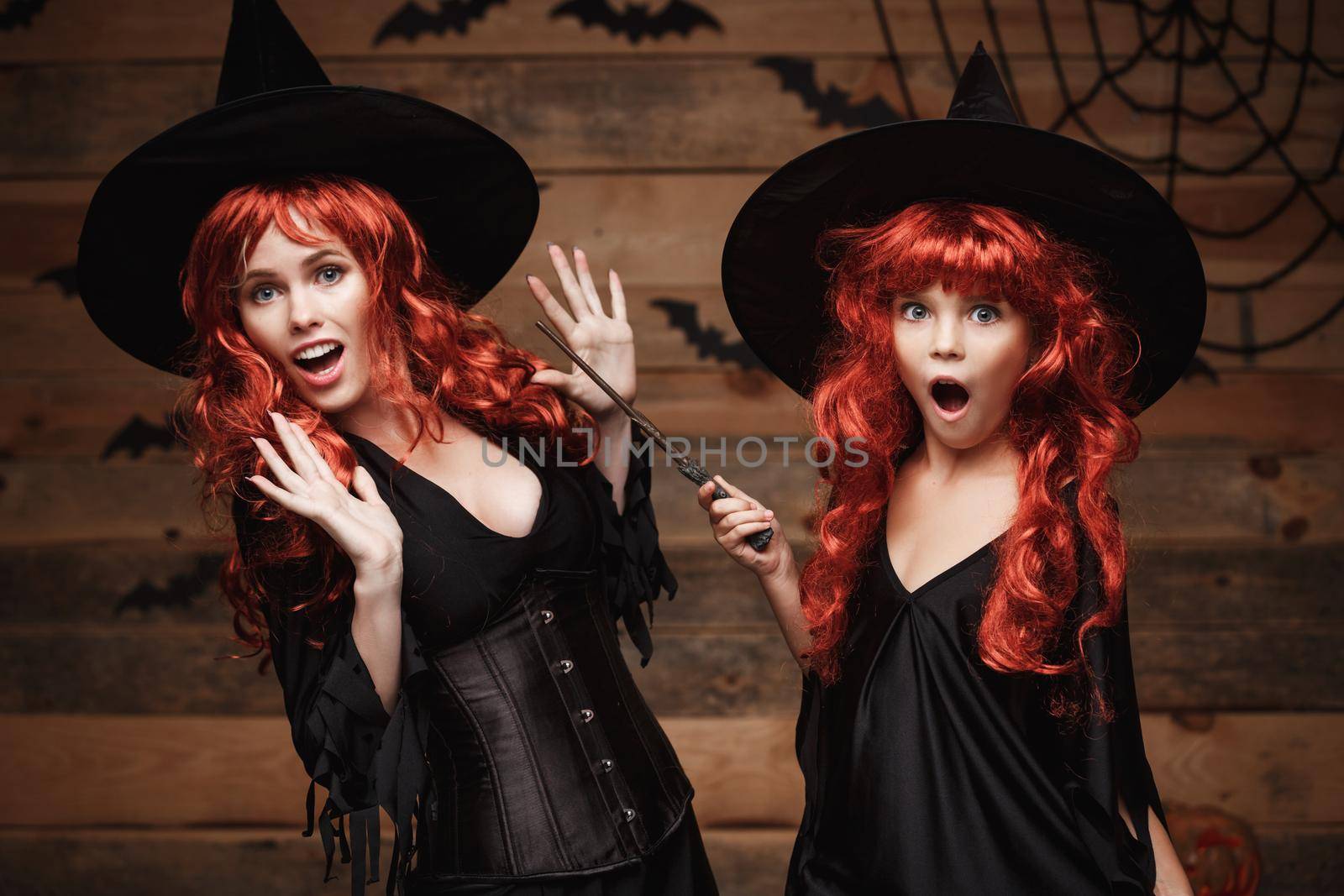 Halloween Concept - Beautiful caucasian mother and her daughter with long red hair in witch costumes and magic wand celebrating Halloween posing with over bats and spider web on Wooden studio background.