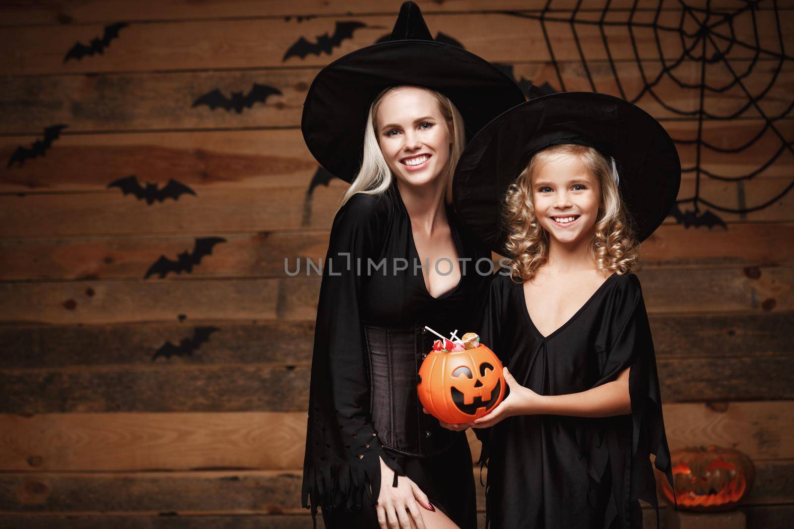 Halloween Concept - beautiful caucasian mother and her daughter in witch costumes celebrating Halloween with sharing Halloween candy and sweet over bats and spider web on Wooden studio background. by Benzoix