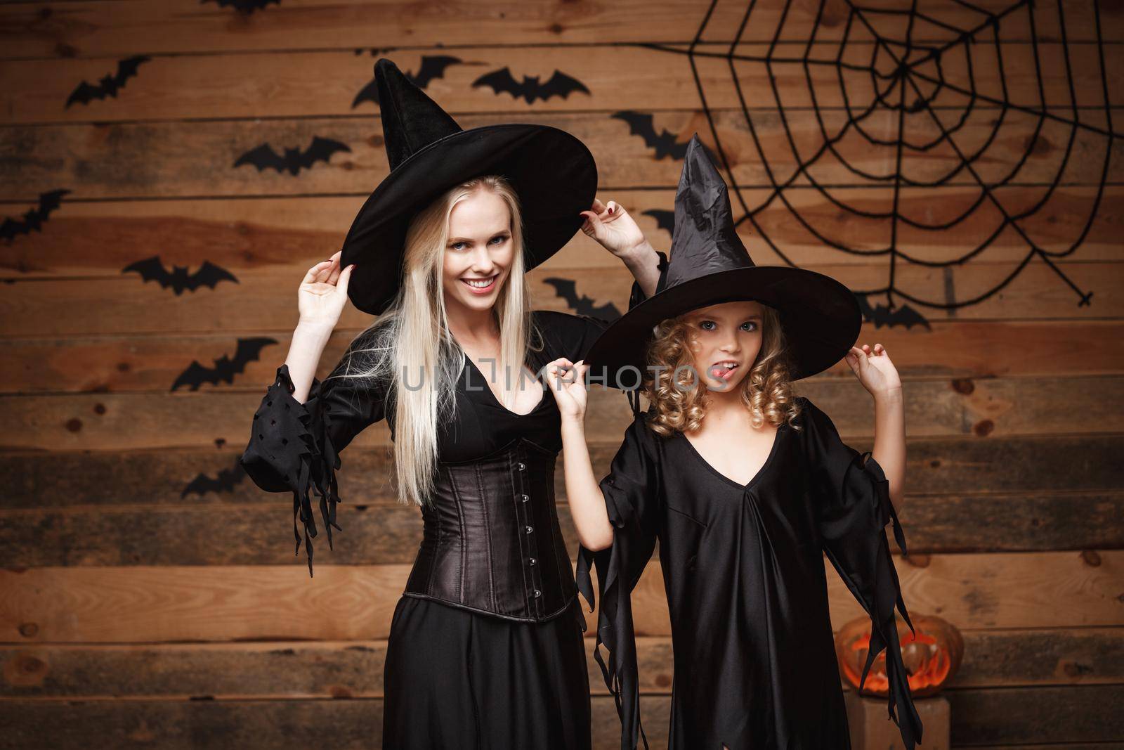Halloween Concept - cheerful mother and her daughter in witch costumes celebrating Halloween posing with curved pumpkins over bats and spider web on Wooden studio background. by Benzoix